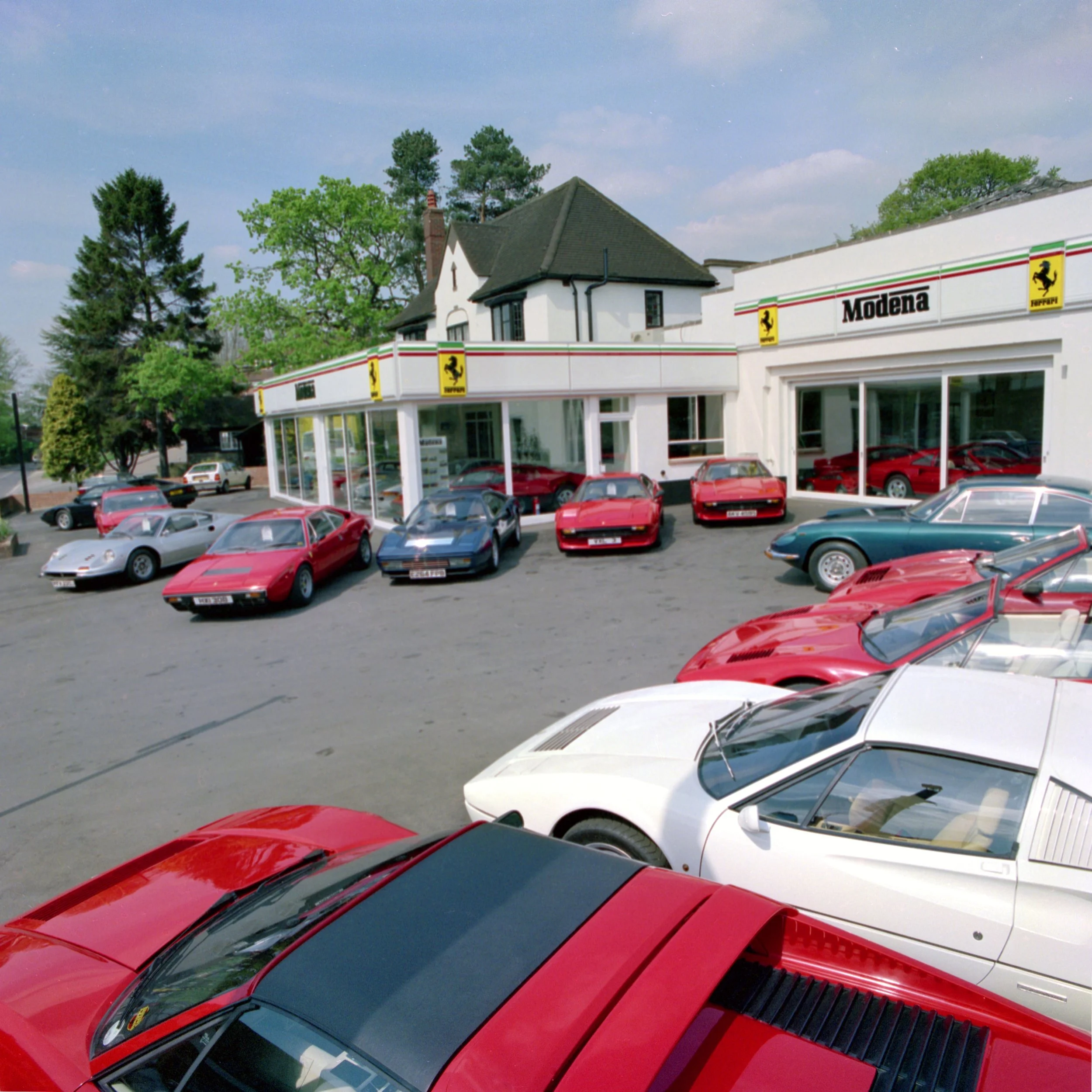 Line of red and white sports cars parked in a lot in front of a building with Ferrari and Modena signs, surrounded by trees and a house with a pitched roof.