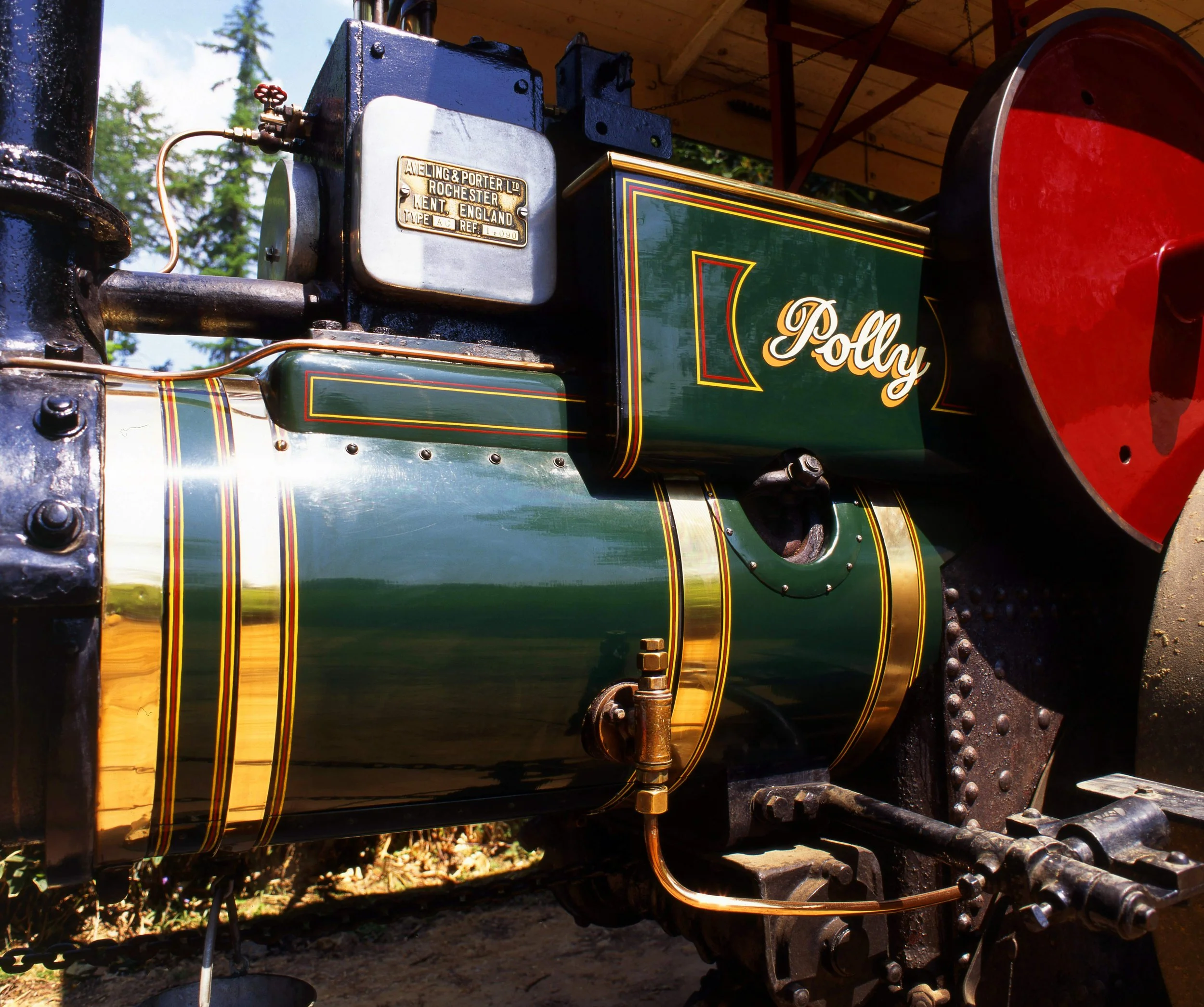 Close-up of a vintage steam locomotive engine with a brass plate, green and black painted body, and red wheels, outdoors under a wooden canopy with trees in the background.