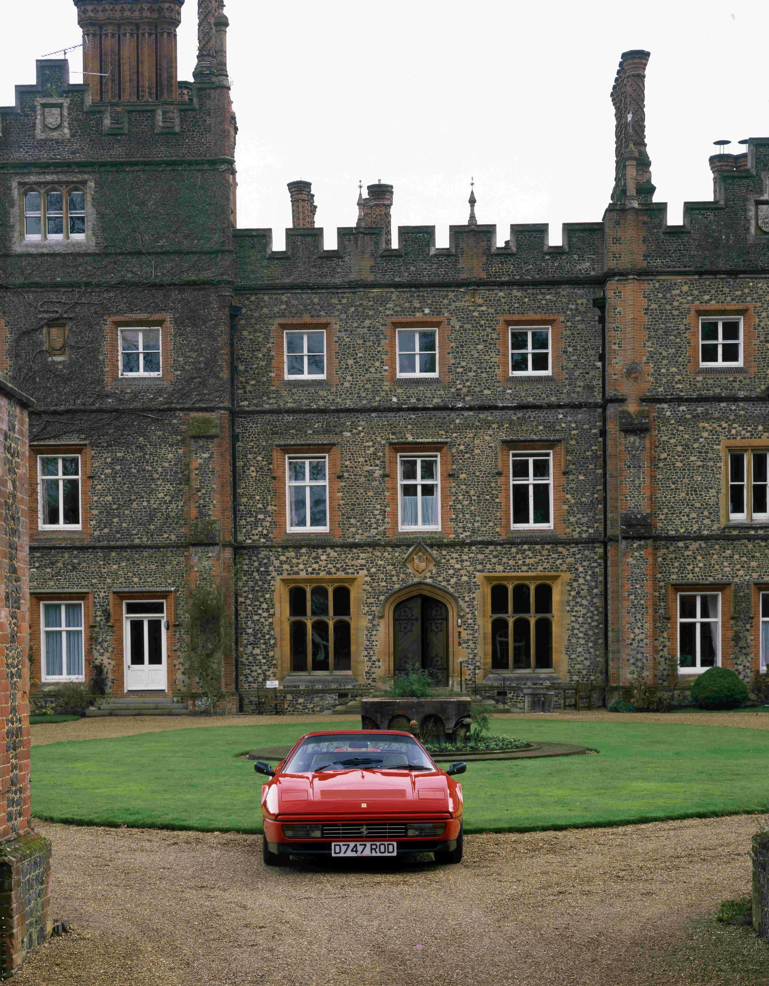 A red Ferrari sports car parked on a gravel driveway in front of a historic castle with a stone exterior, numerous windows, and tall towers with decorated chimney stacks.