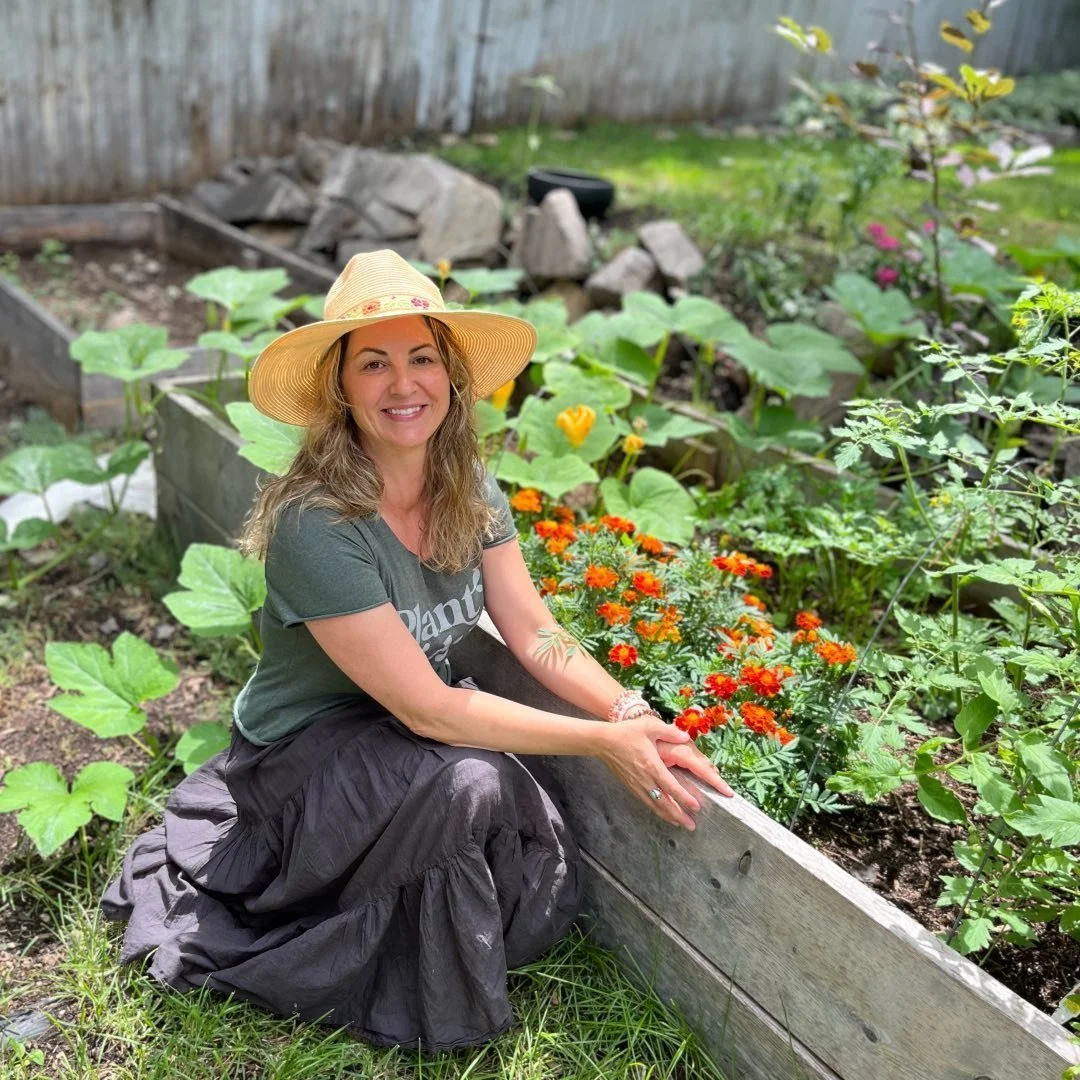 A woman smiling and sitting on the edge of a garden bed with orange and yellow flowers, wearing a wide-brimmed straw hat, green t-shirt, and black skirt, surrounded by green plants and a wooden fence in the background.