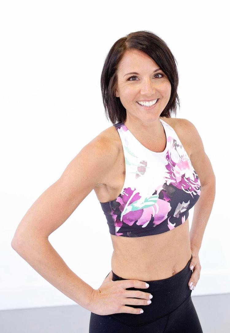 A woman with short dark hair smiling, wearing a floral sports bra and black workout leggings, standing with hands on her hips against a plain white background.