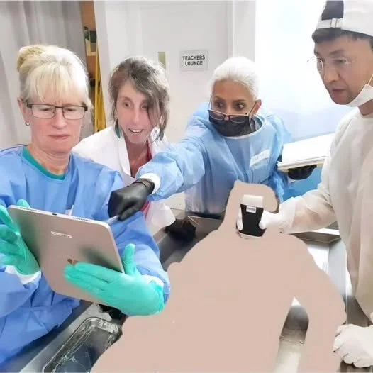 Medical professionals wearing scrubs and gloves gathered around a table, looking at a tablet, with a silhouette of a human body model on the table.