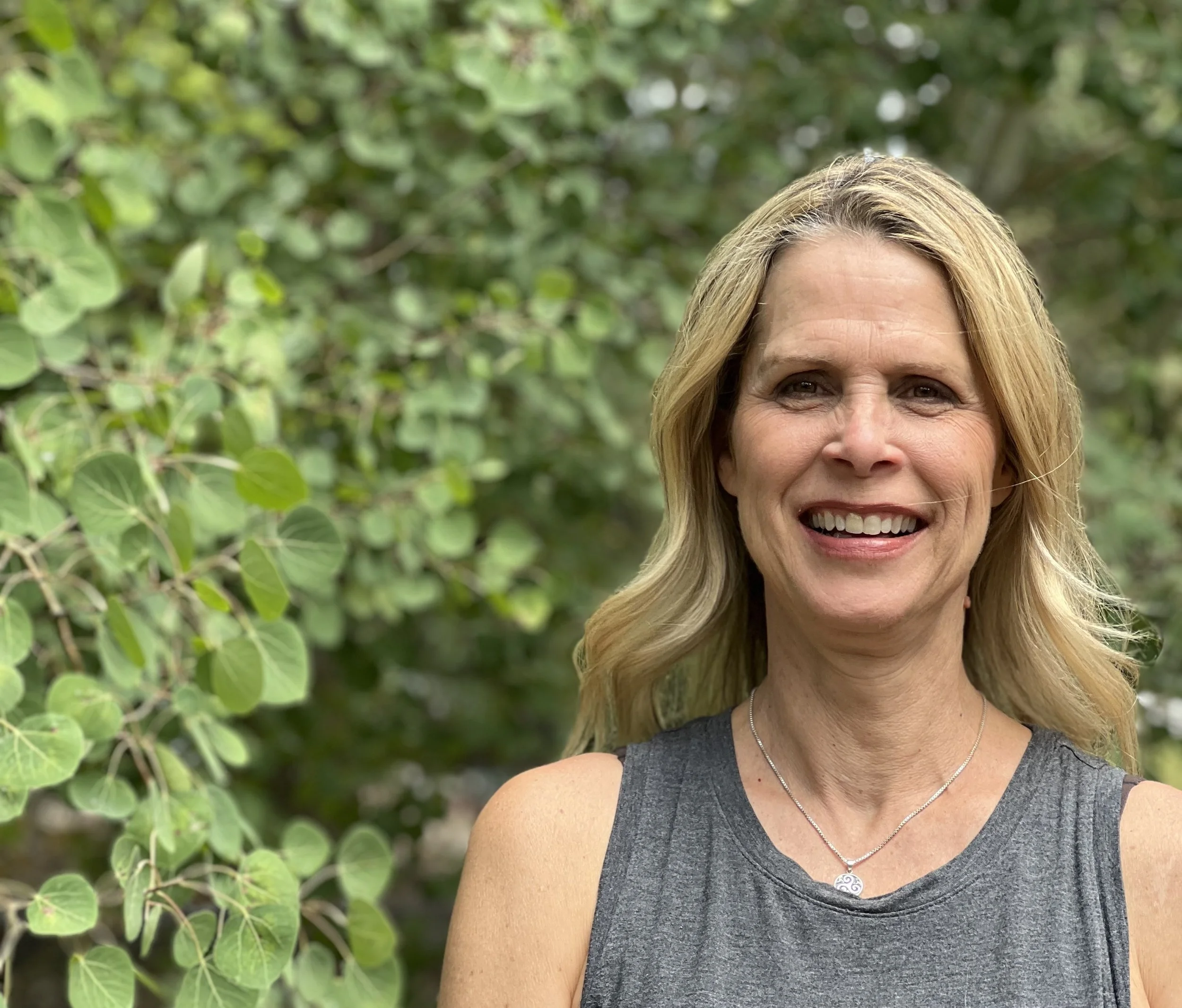 A woman with blonde hair, wearing a sleeveless gray top and a necklace, smiling outdoors in front of green foliage.