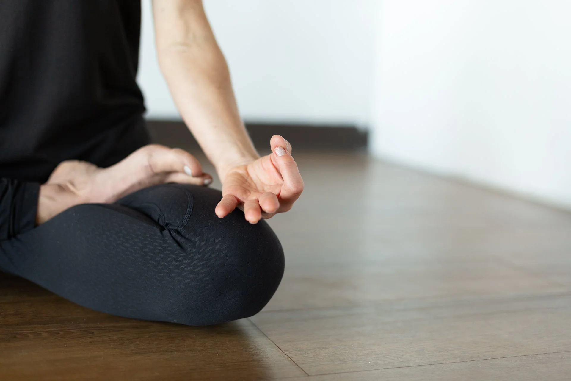 A person practicing yoga or meditation, sitting cross-legged on a wooden floor, with one hand resting on their knee with fingers forming a mudra, wearing black athletic clothing.