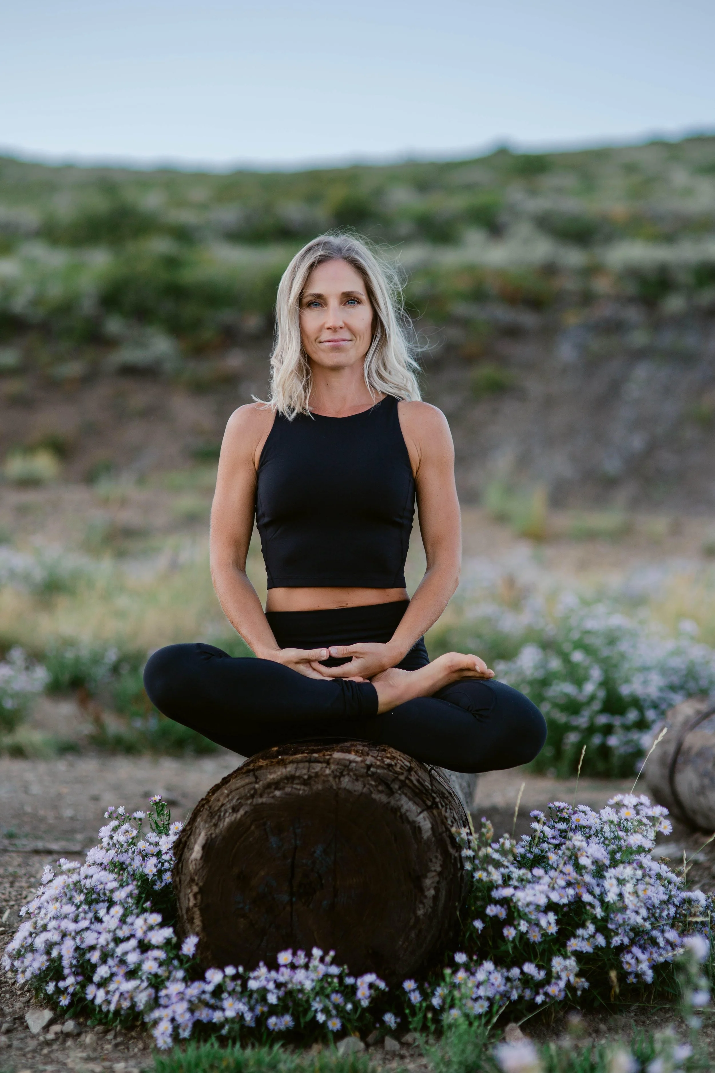 A woman with blonde hair, wearing a black sleeveless top and leggings, sitting cross-legged on a tree trunk in an outdoor natural setting with green hills and wildflowers.