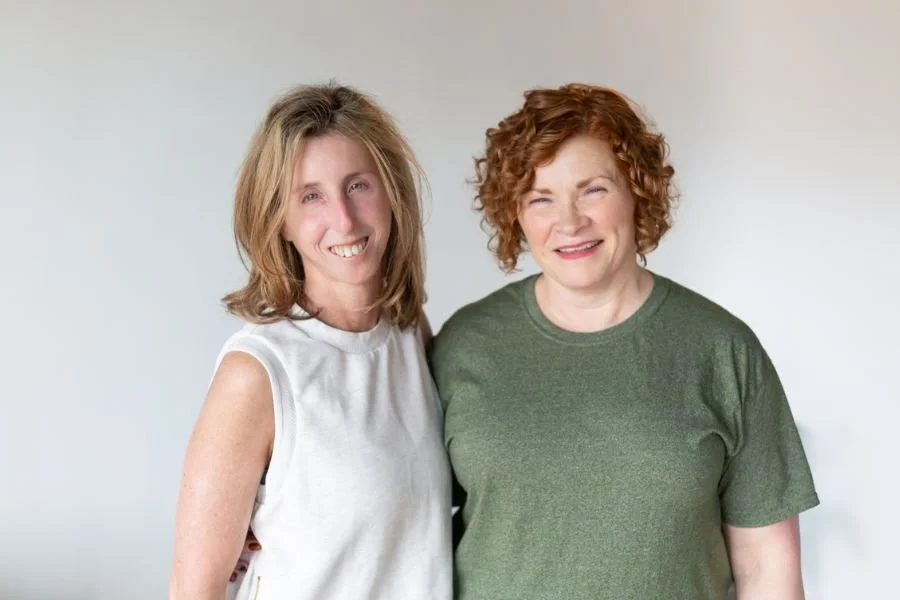 Two women smiling and standing close together against a white background.