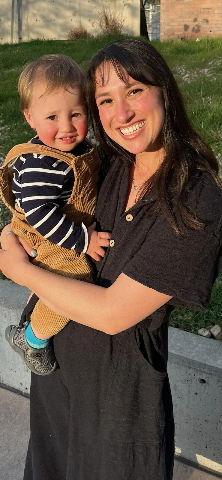 A smiling woman wearing a green cap with sunglasses on top and a young boy with blond hair and a gray shirt with colorful stripes on the chest, are outdoors in a lush, green forest. In the background, there is a black and tan dog behind dense bushes and trees.