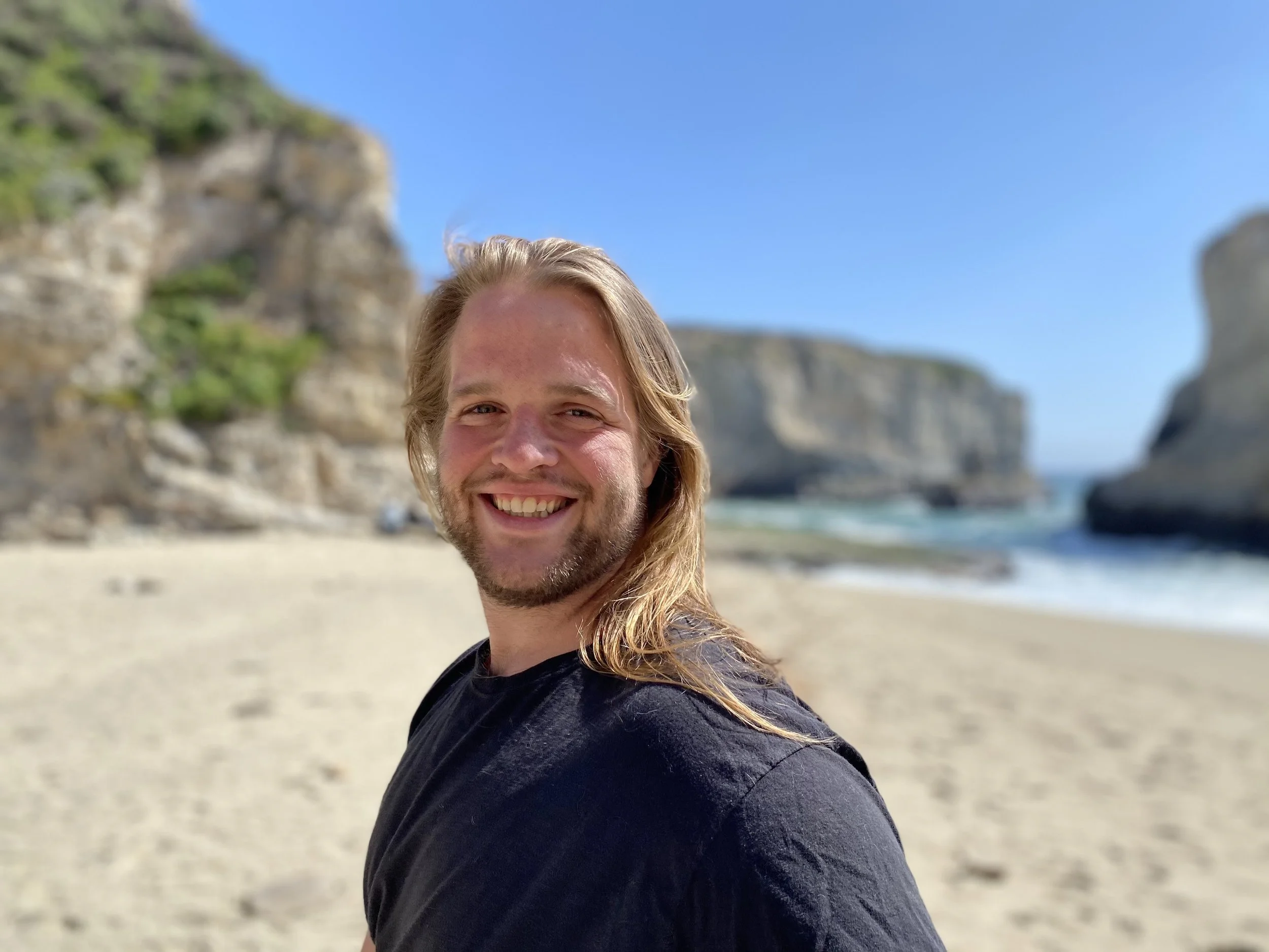 A smiling man with long blond hair and a beard at the beach with cliffs and blue sky in the background.