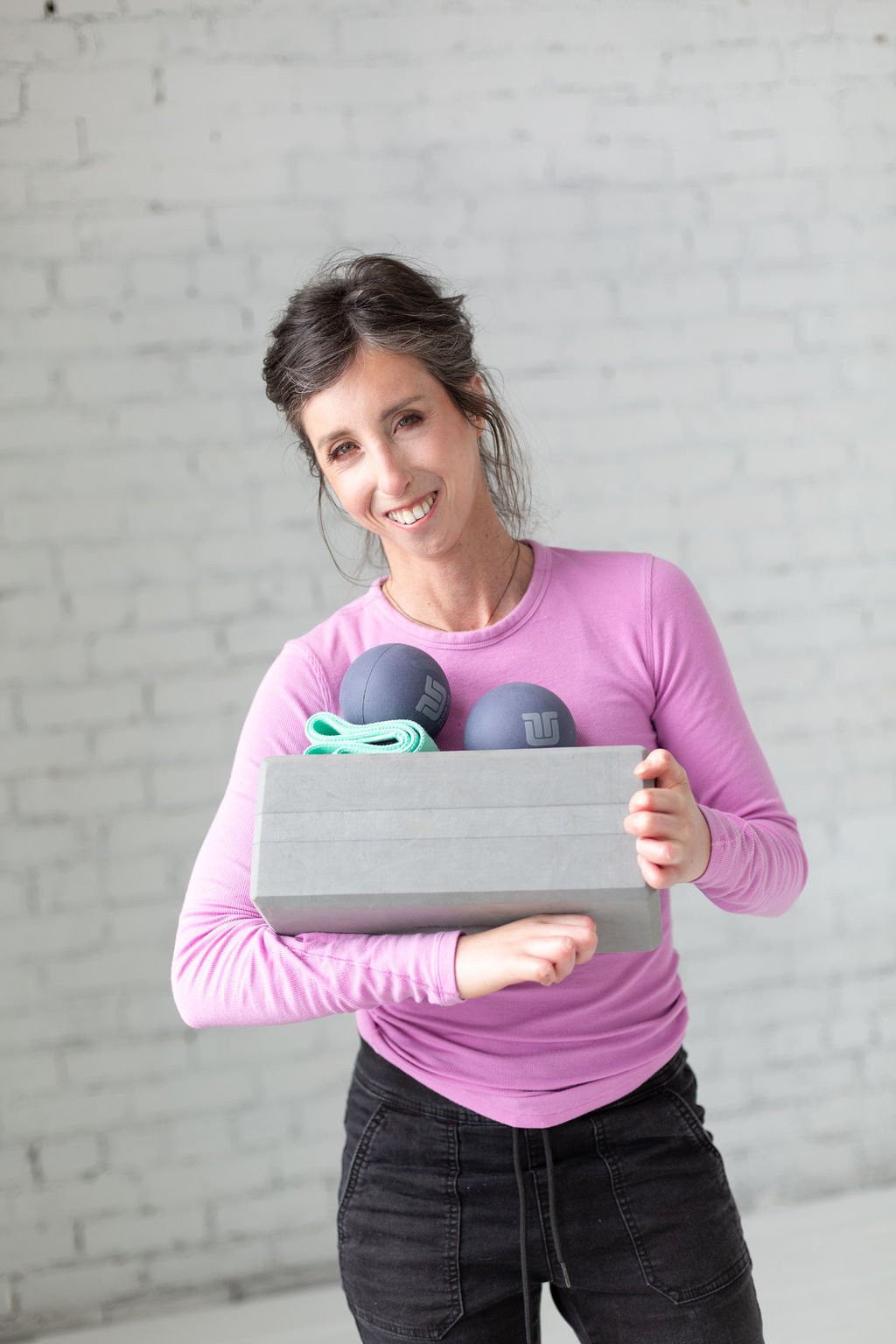 Woman with dark hair in a ponytail, wearing a pink long sleeve shirt, smiling and holding a gray box with two black stress balls and green shoelace inside, standing in front of a white brick wall.