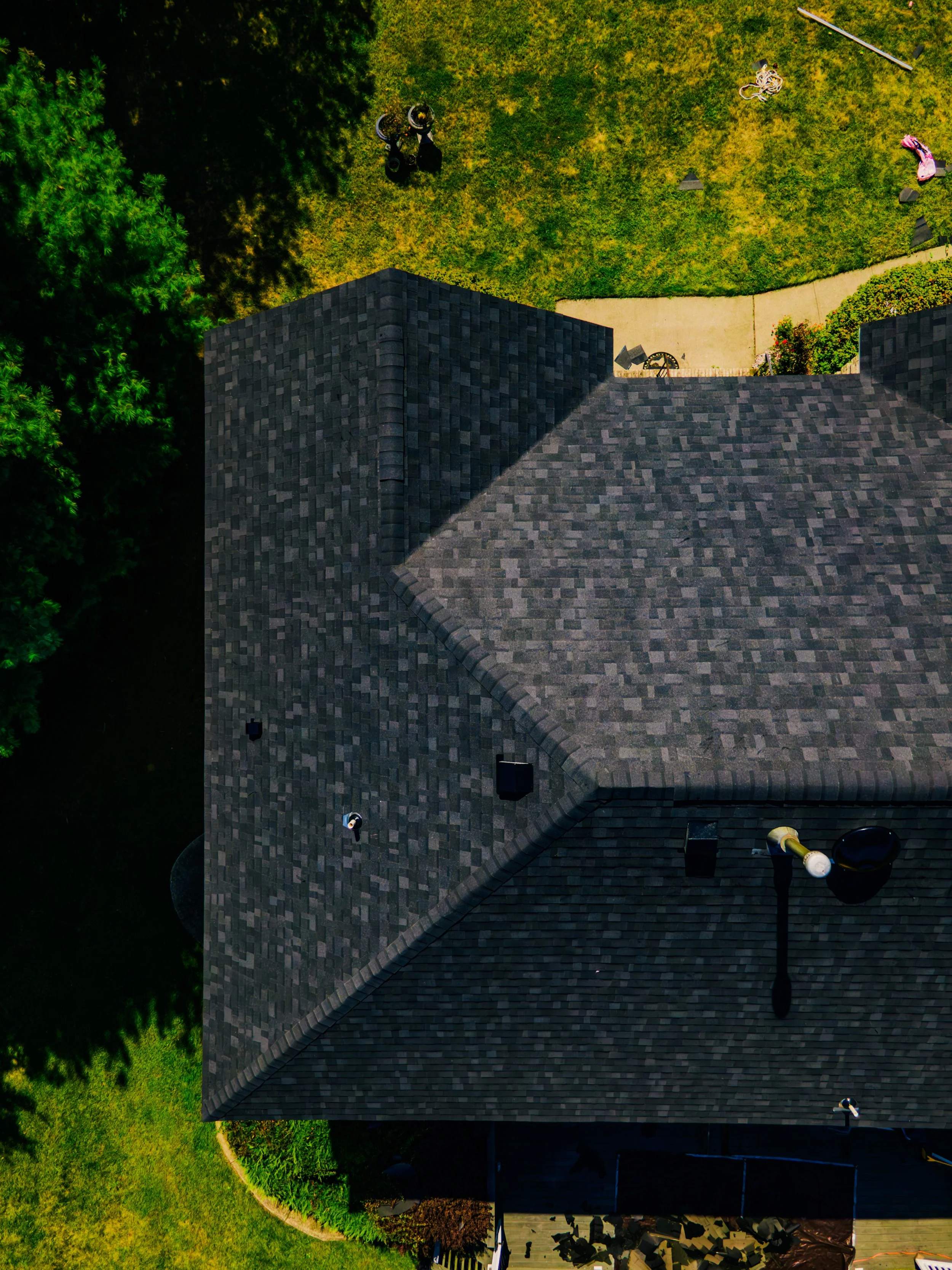 Aerial view of a house with a dark shingled roof, trees, and a backyard with a walkway, lawn, and outdoor furniture.