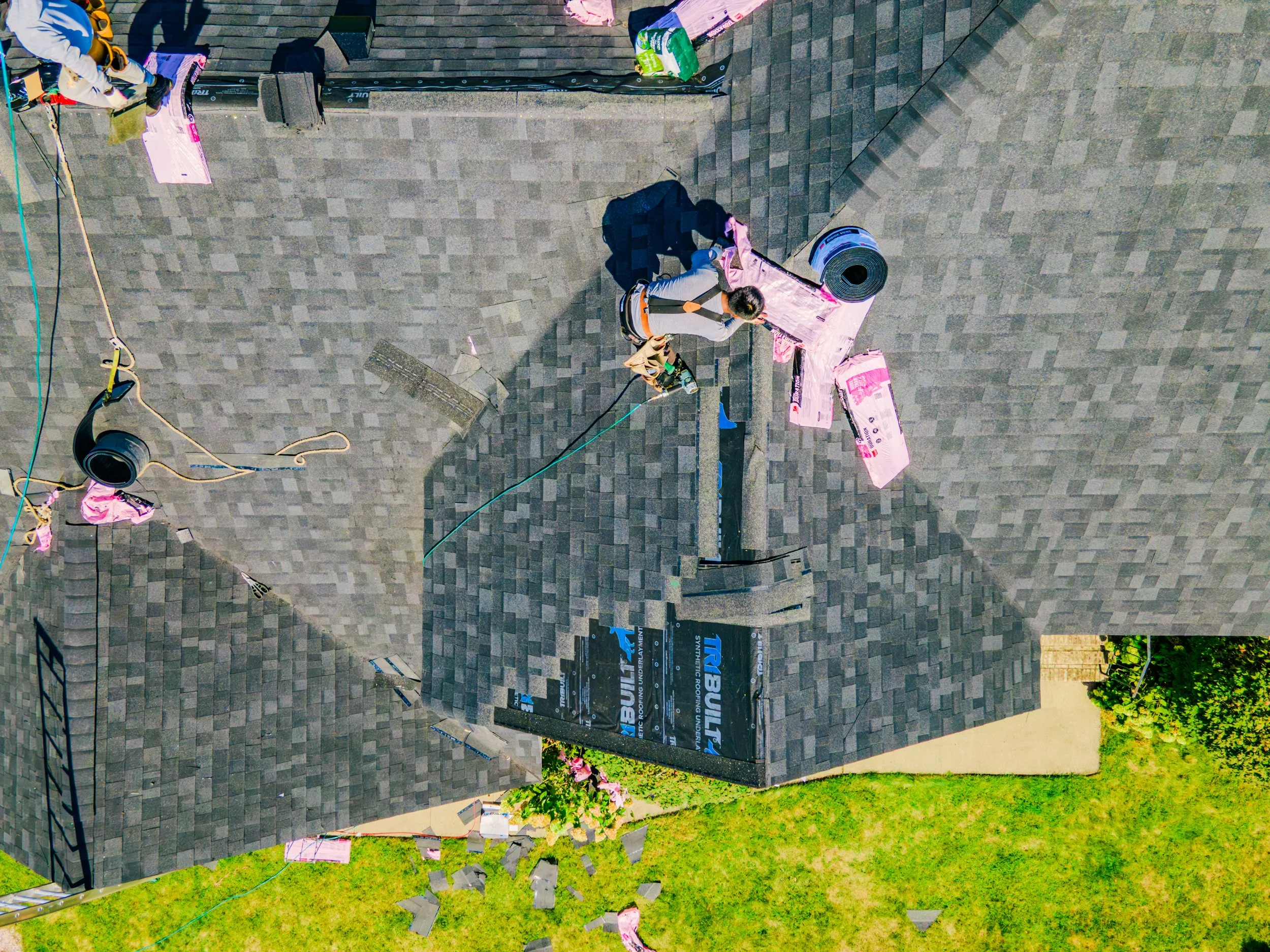 Aerial view of a house roof under construction with workers installing shingles, roofing tools, and materials, surrounded by green grass.
