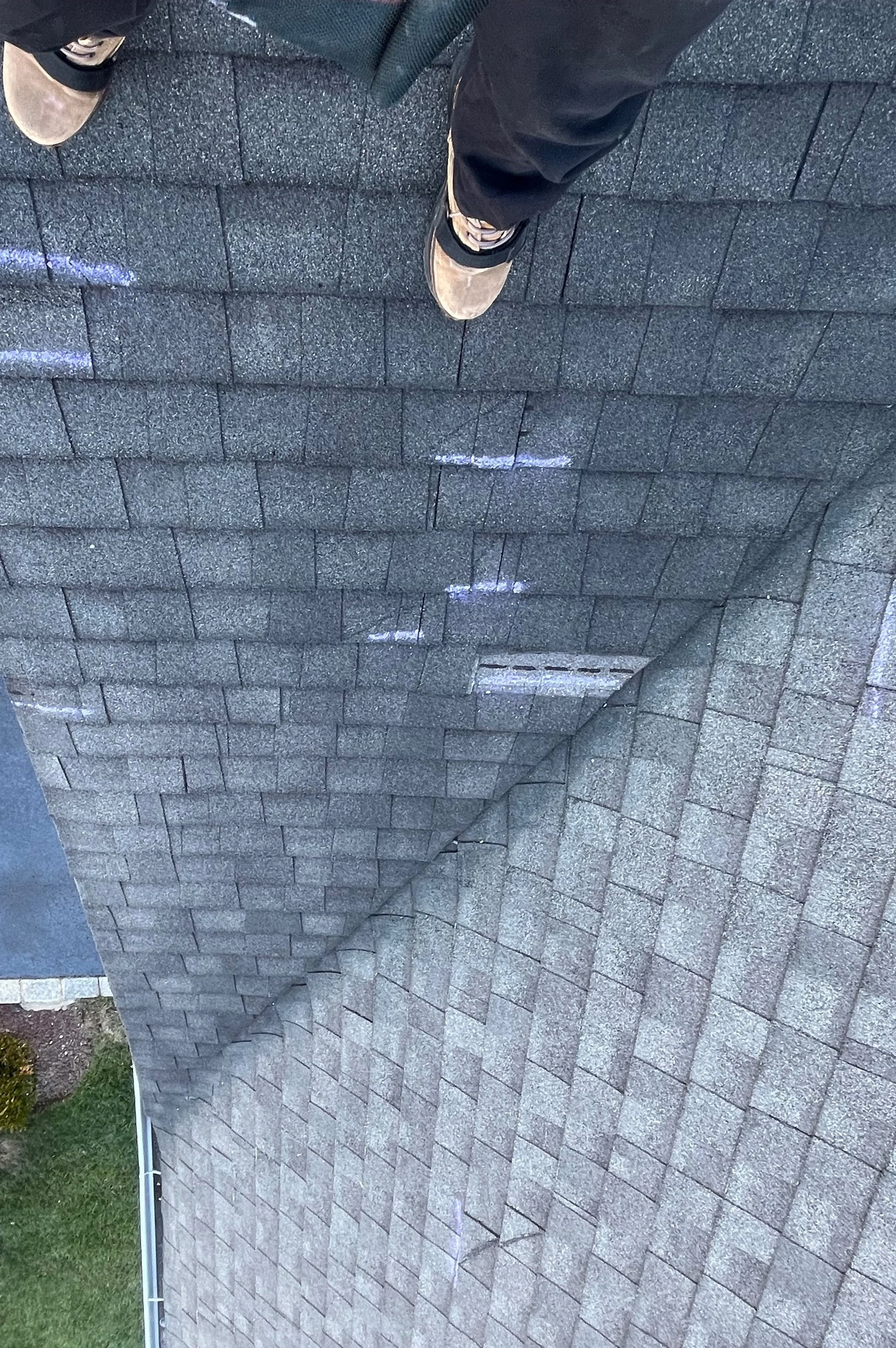 View of a rooftop with grey shingles and a person's feet visible, standing on the roof. The person is wearing sneakers and dark pants. Green grass and shrubbery are visible in the lower left corner.