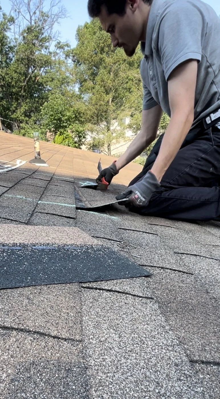 A man kneeling on a roof installing or repairing asphalt shingles, using tools, with trees and a clear sky in the background.