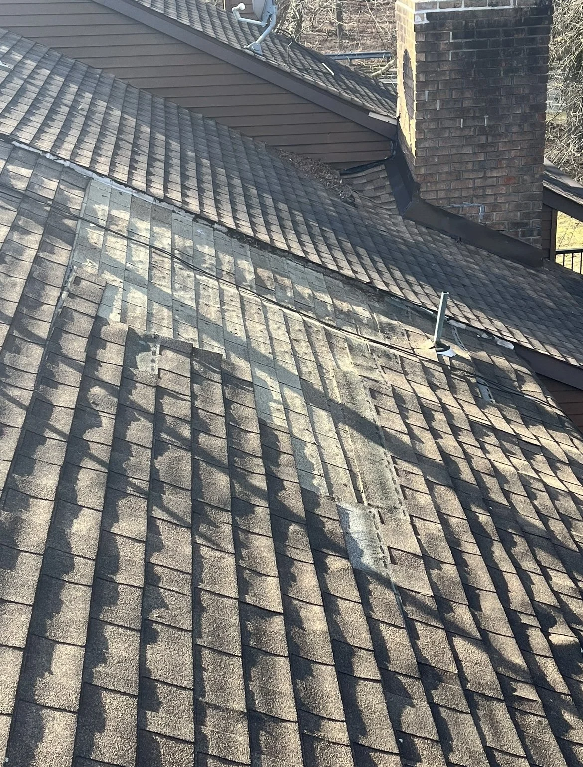 View of a sloped roof with asphalt shingles, some parts appear weathered and worn, with a metal pipe protruding from the roof near the chimney, which is made of bricks.