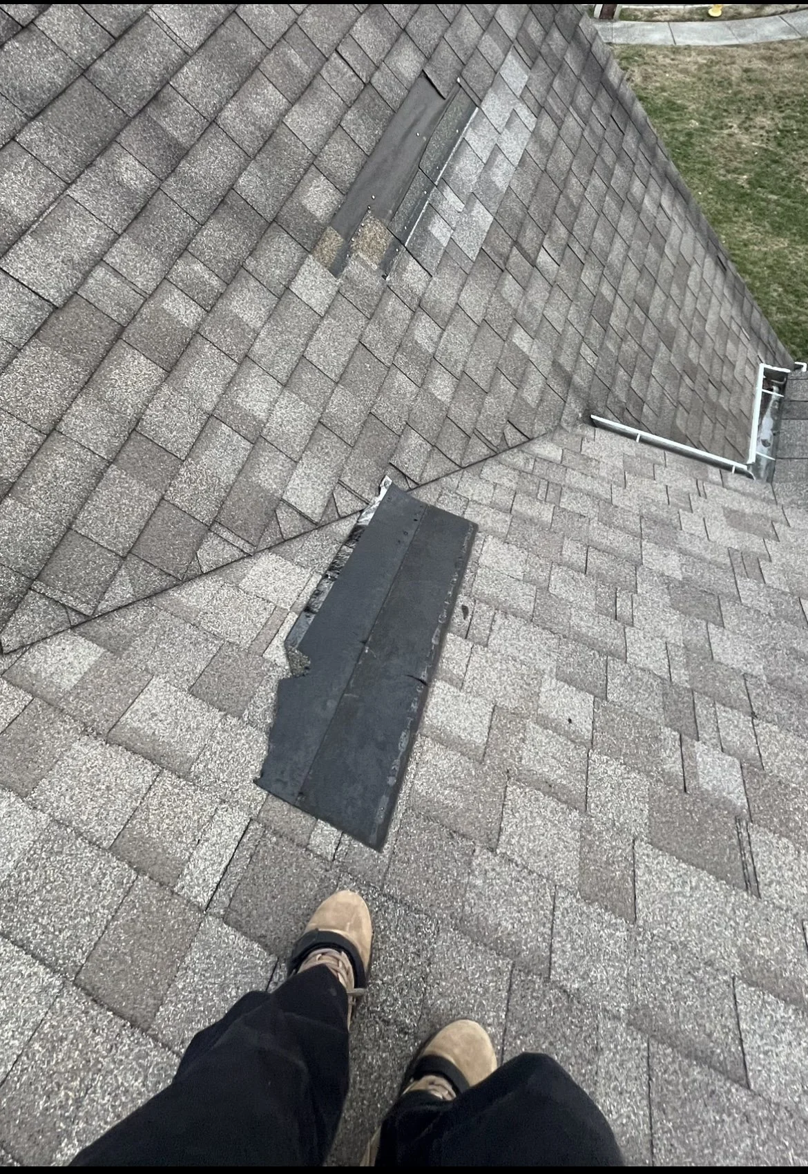 View of a shingled roof with a section under repair, showing a black patch and a person's feet at the edge of the roof, with grass visible in the background.