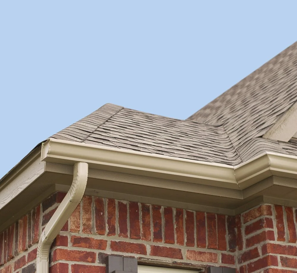 Close-up of a house corner showing roof shingles, gutter, and brick wall with window.