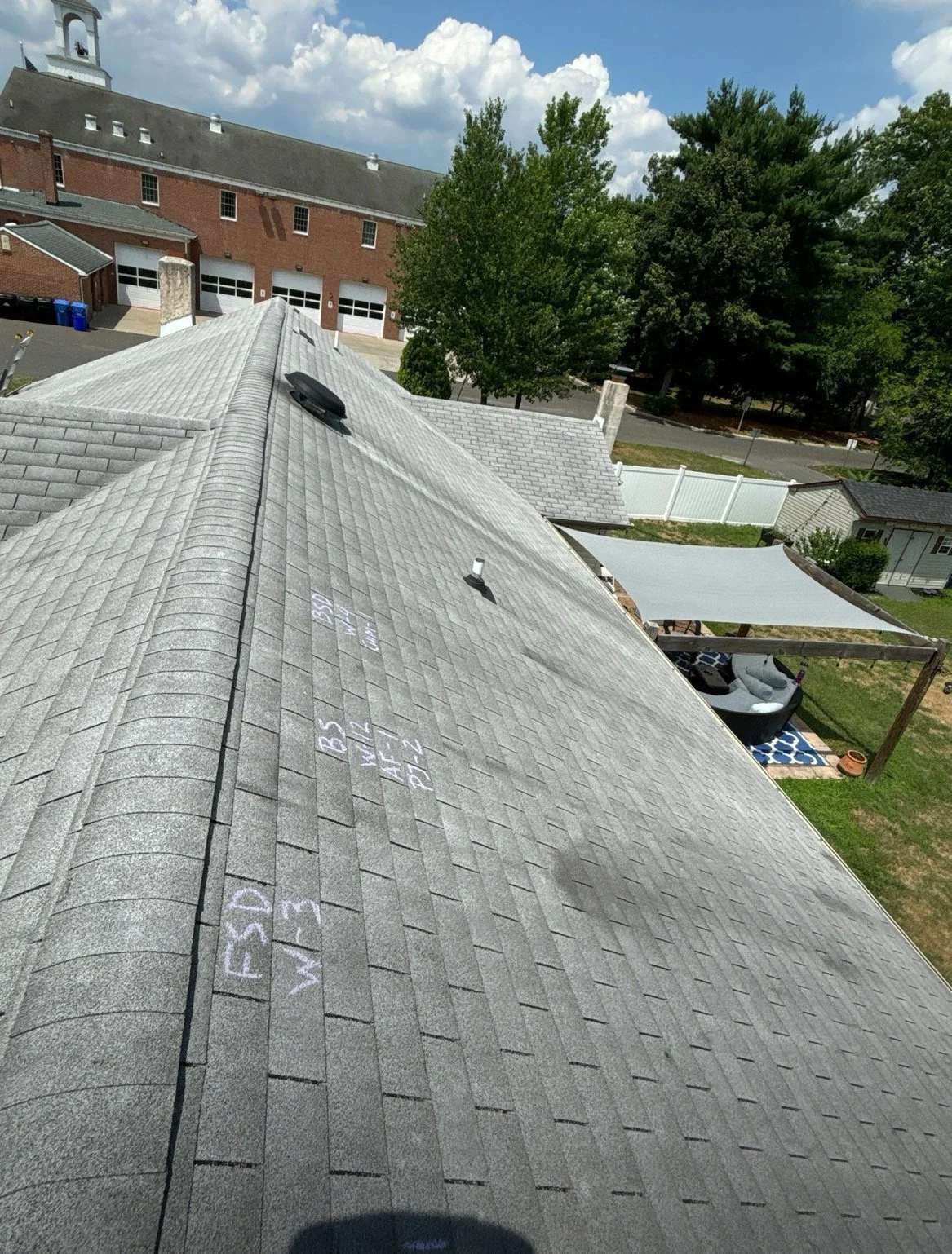 View of a house roof with visible shingles, skylights, and roofing chalk markings. A backyard with patio furniture, a shade sail, and trees is visible in the background.
