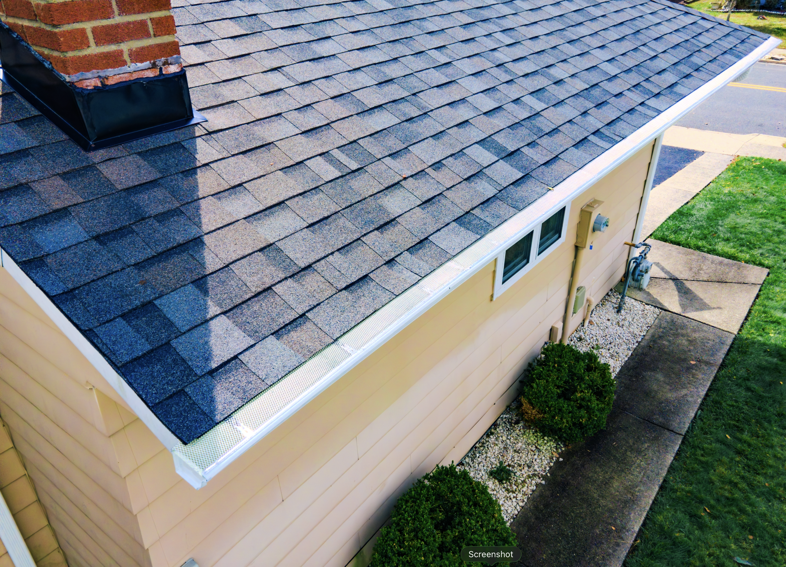 Aerial view of a house's roof with shingles, surrounding sidewalk, and nearby green lawn and bushes.