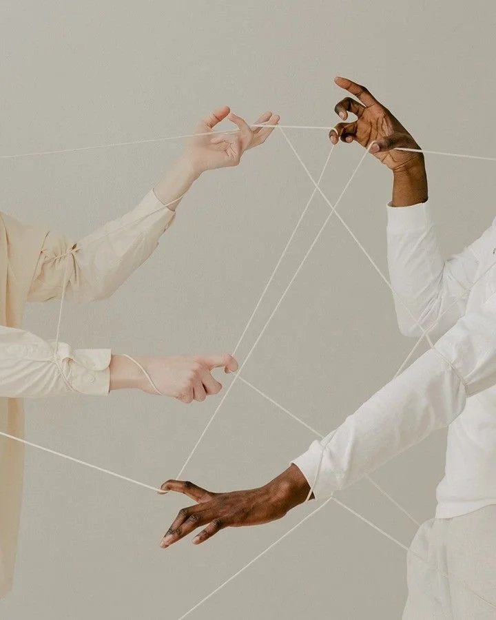 Multiple hands of different skin tones are intertwined with white strings, creating a web-like structure against a neutral background.