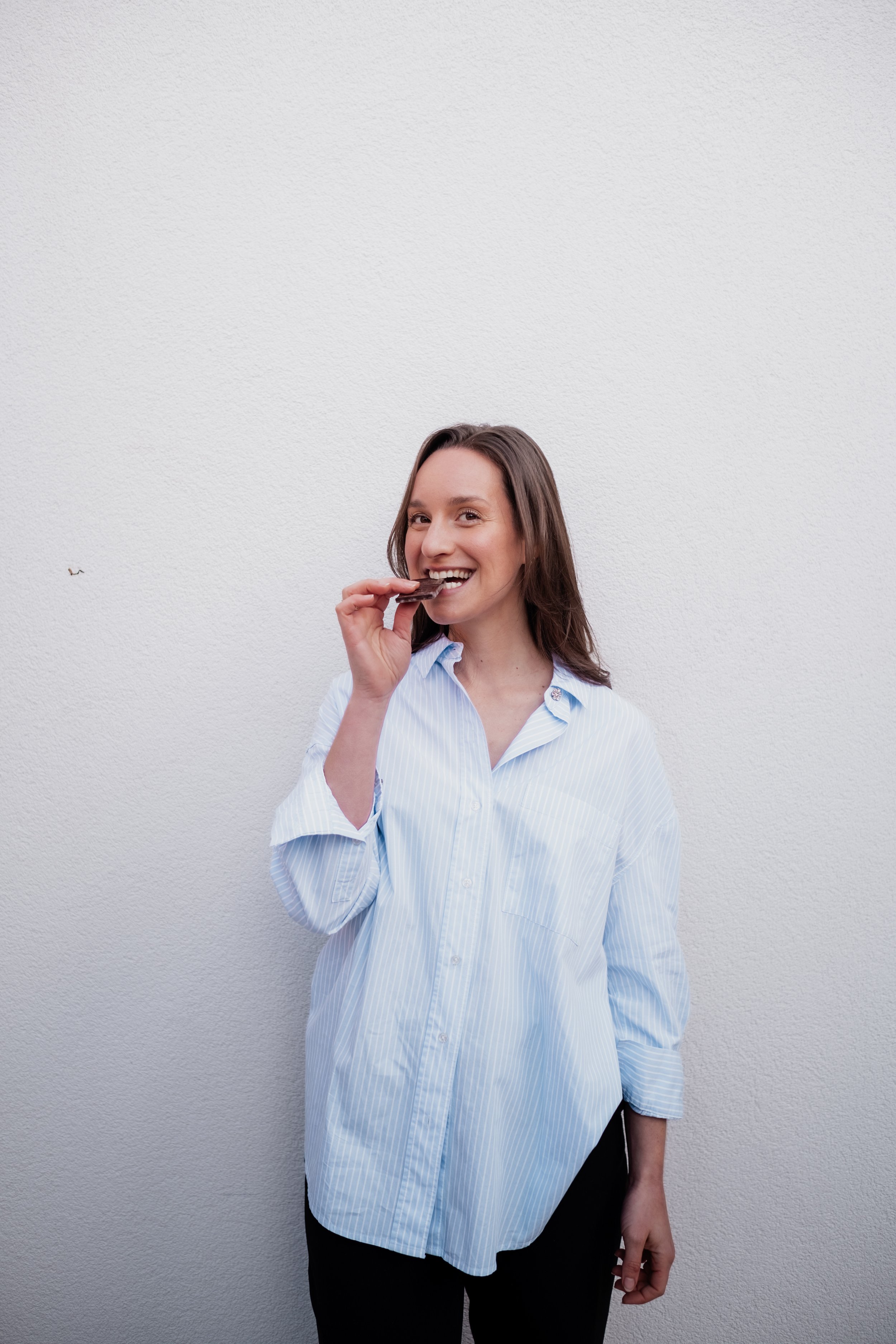 A woman with brown hair smiling and eating a chocolate bar standing against a white wall.
