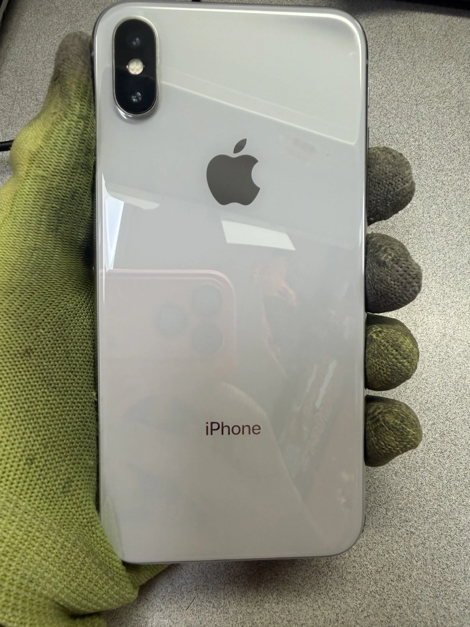 Back view of a white iPhone with dual camera, held by a hand wearing a yellow-green knitted glove, resting on a beige surface.