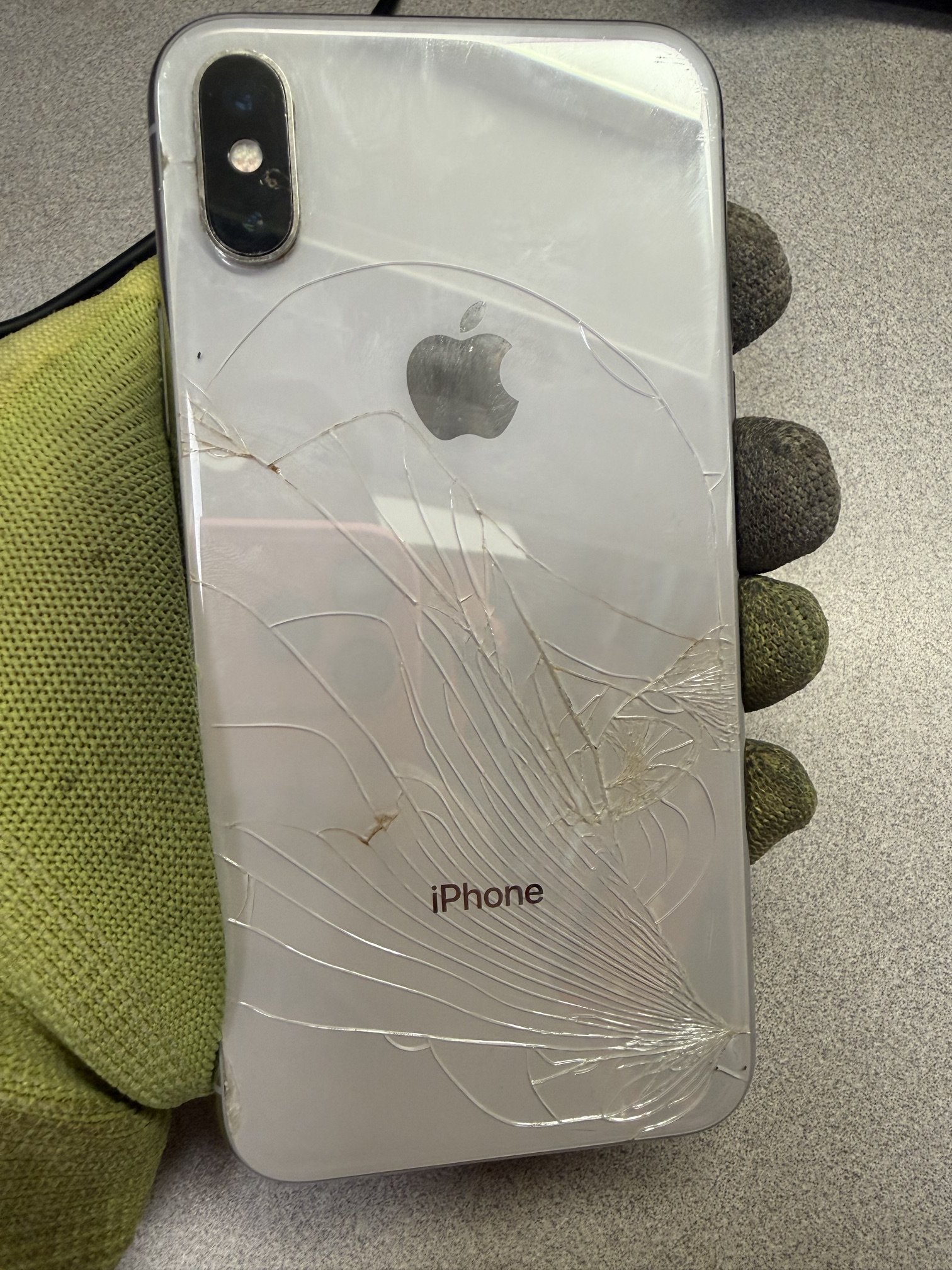 Cracked silver iPhone with a visible Apple logo and triple camera lens, resting on a speckled gray surface with a green textured cloth partially visible.