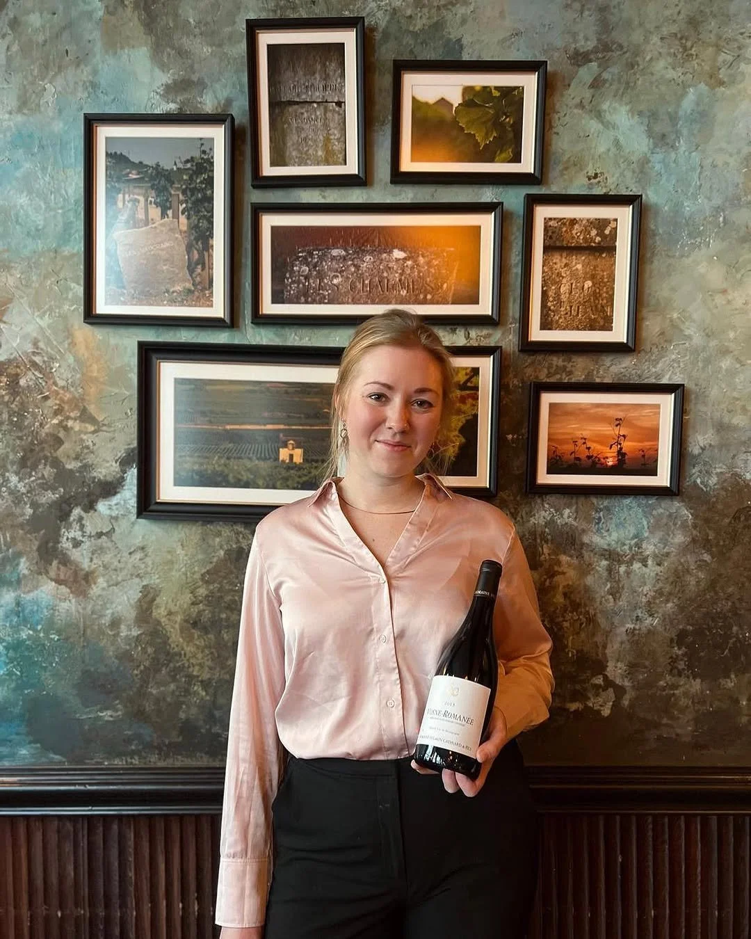 A woman in a satin pink blouse holding a bottle of red wine. She is standing in front of a textured wall with a gallery of framed photographs, including vineyards and landscapes.