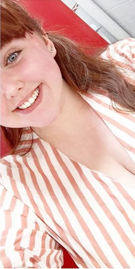 A young woman with long red hair and blue eyes, smiling, wearing a striped pink and white blouse, sitting in an indoor setting with red and white walls.