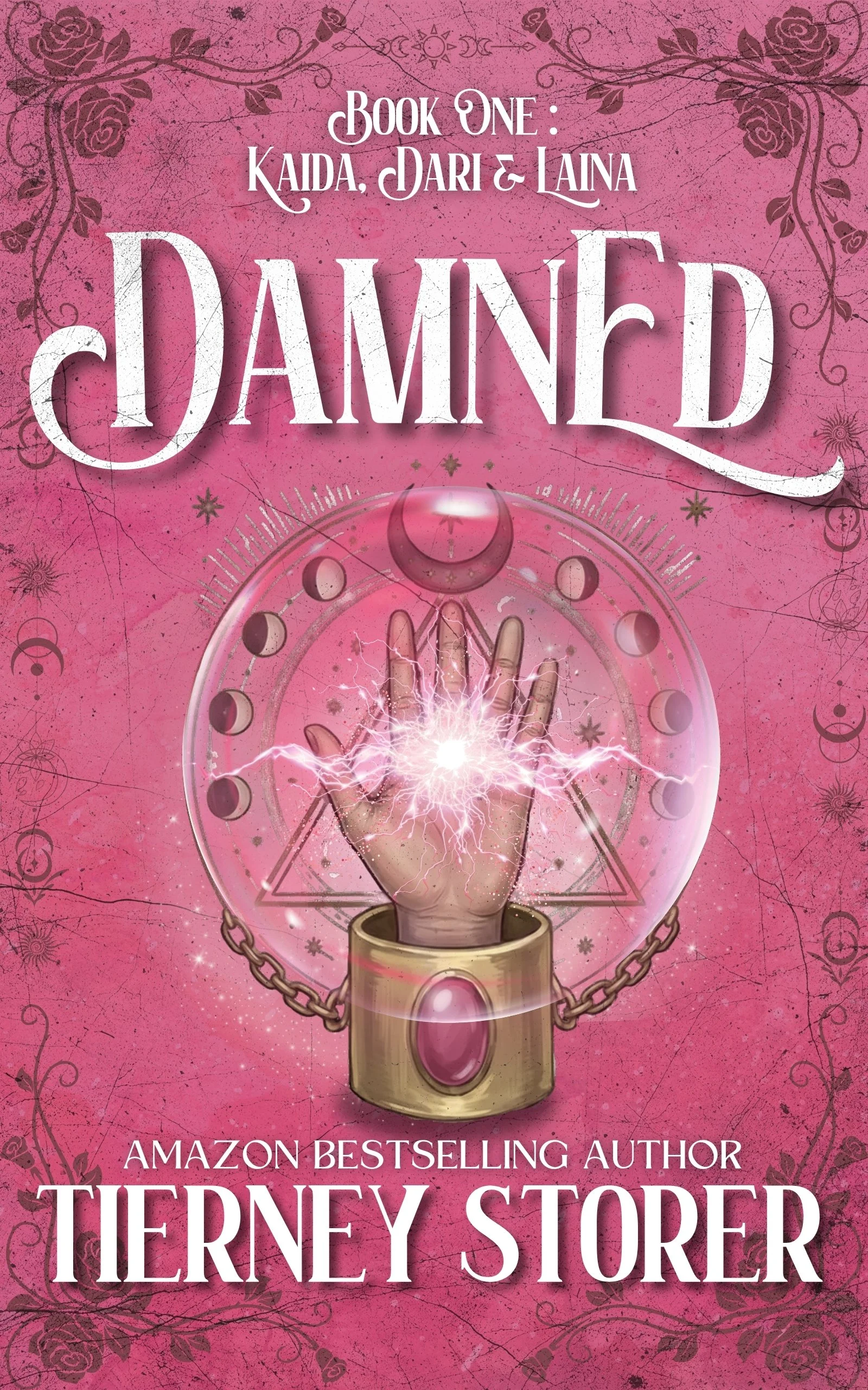 The book cover is pink with black floral and swirl designs in the corners. It features a mystical illustration of a hand with lightning emerging from the palm, inside a pentagram and a circular border. The title reads "Dammed" in large white letters. Above it, the text says "Book One: Kaida, Dari & Iaina." Below the title, the author is noted as an "Amazon Bestselling Author," Tremney Storer.