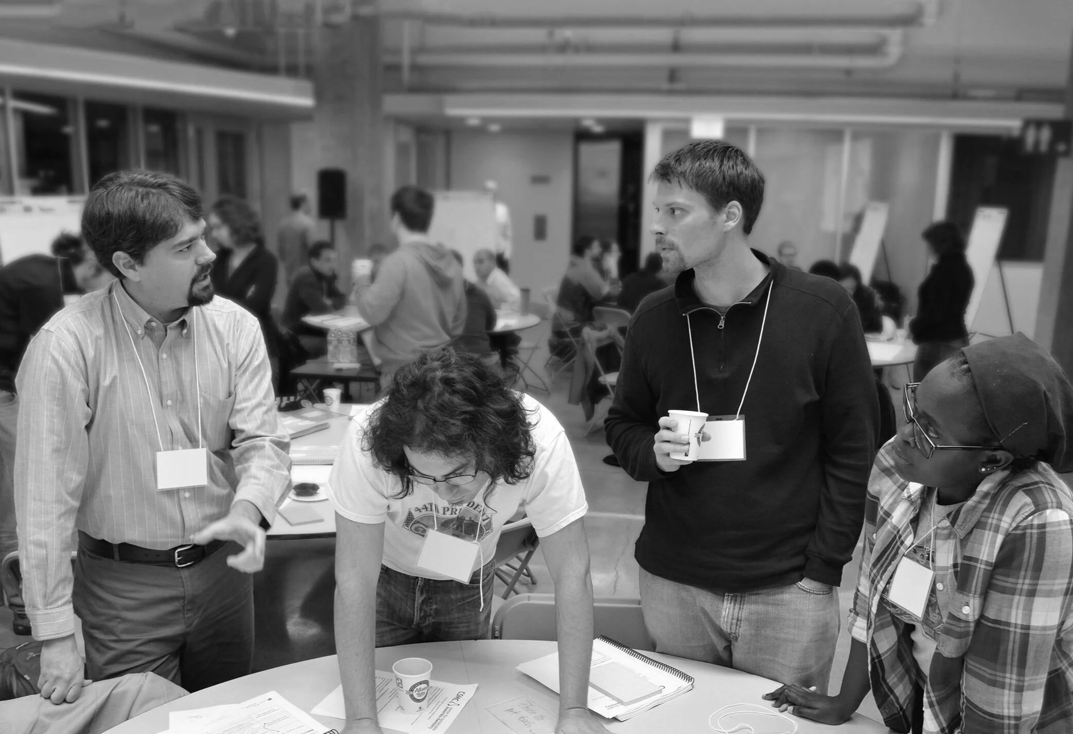 A group of people engaged in a discussion at a conference or workshop in a modern room with tables and presentation boards in the background, some wearing name tags and lanyards.