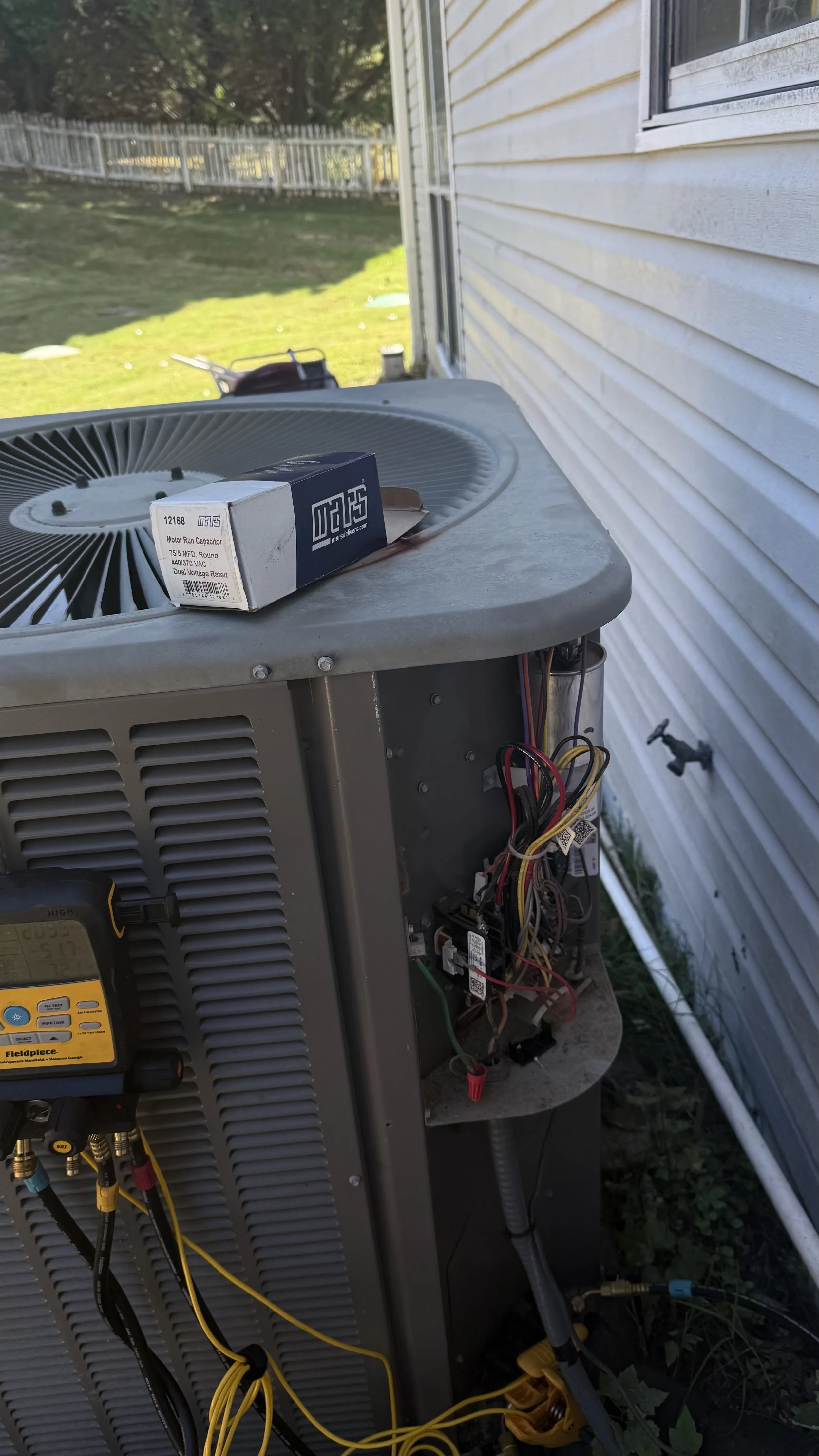 An outdoor air conditioning unit with wires and components exposed, placed beside a house with siding, with a yard and fence in the background.