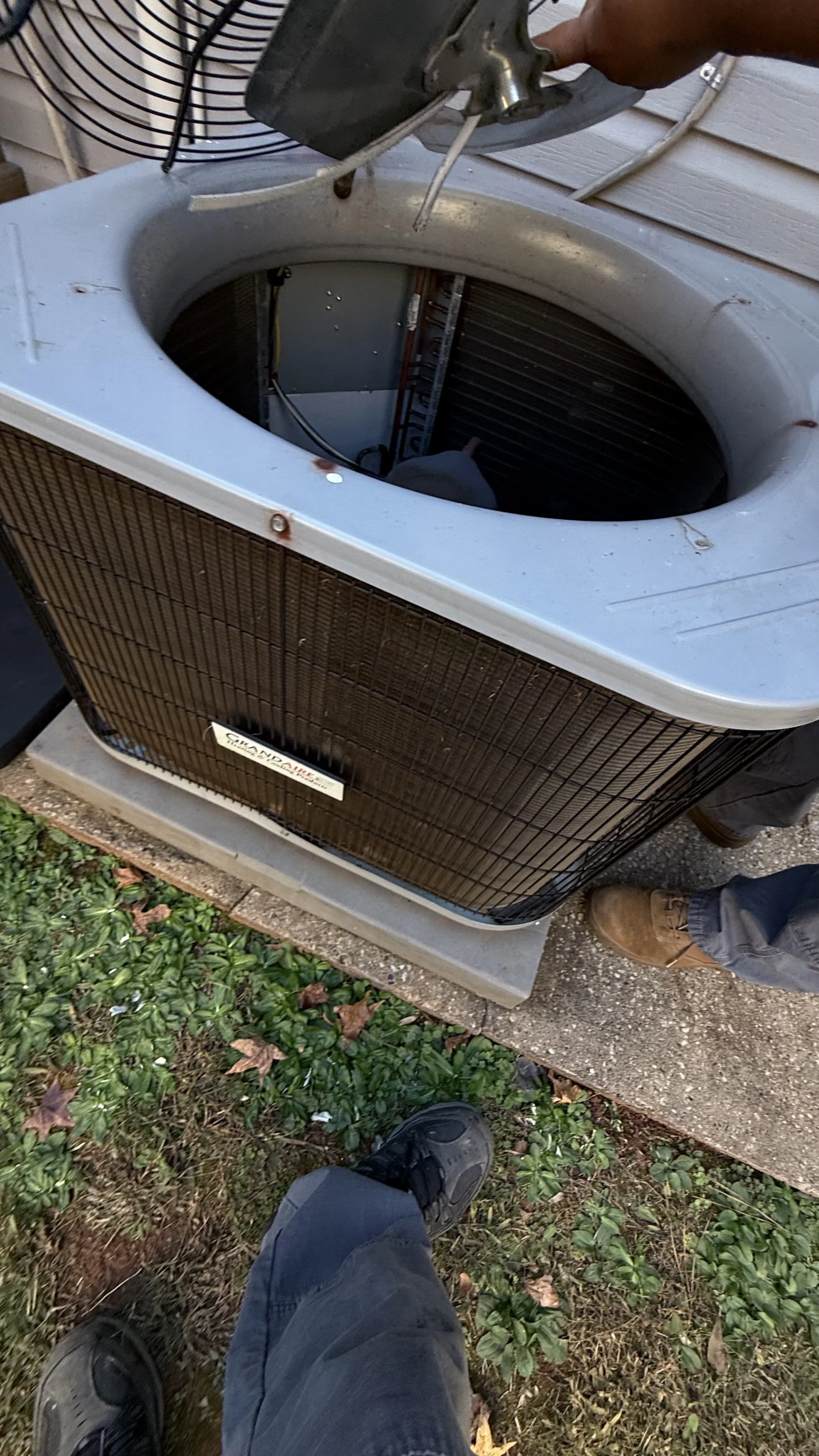 An outdoor air conditioning unit with the top cover removed, revealing the internal components, and two people working on it.