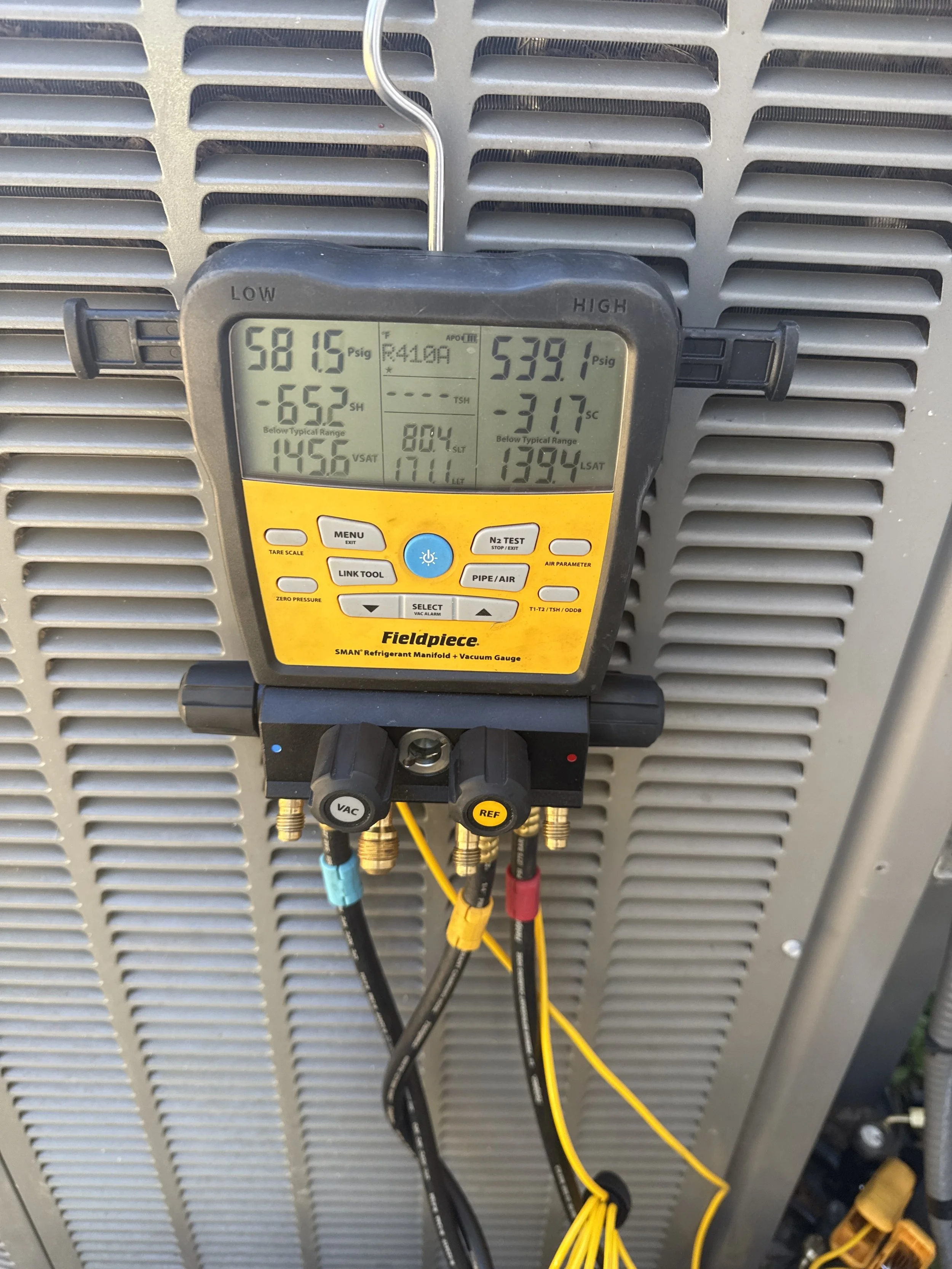 A digital HVAC manifold gauge connected to a unit, displaying pressure readings for refrigerant R410A, with hoses attached and clamped to a grey metal surface.