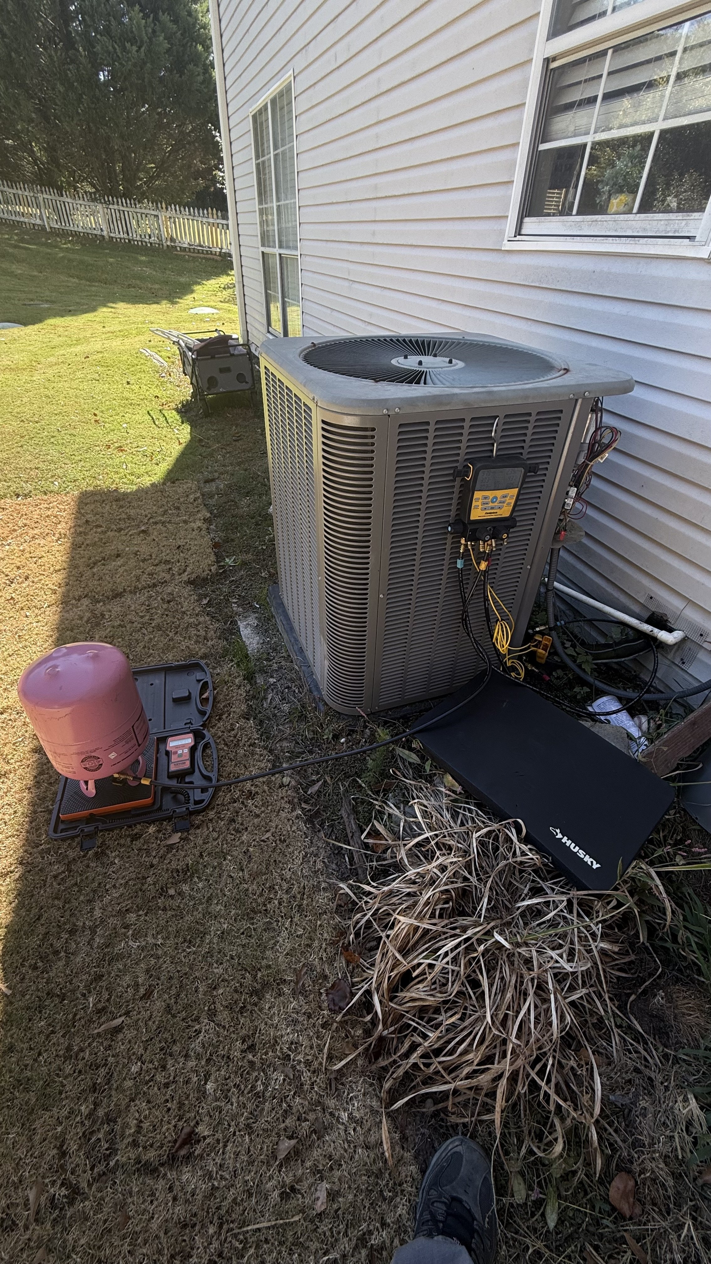 An outdoor view of an air conditioning unit working on the side of a house with tools and a pink refrigerant tank nearby on the lawn, and a white picket fence in the background.