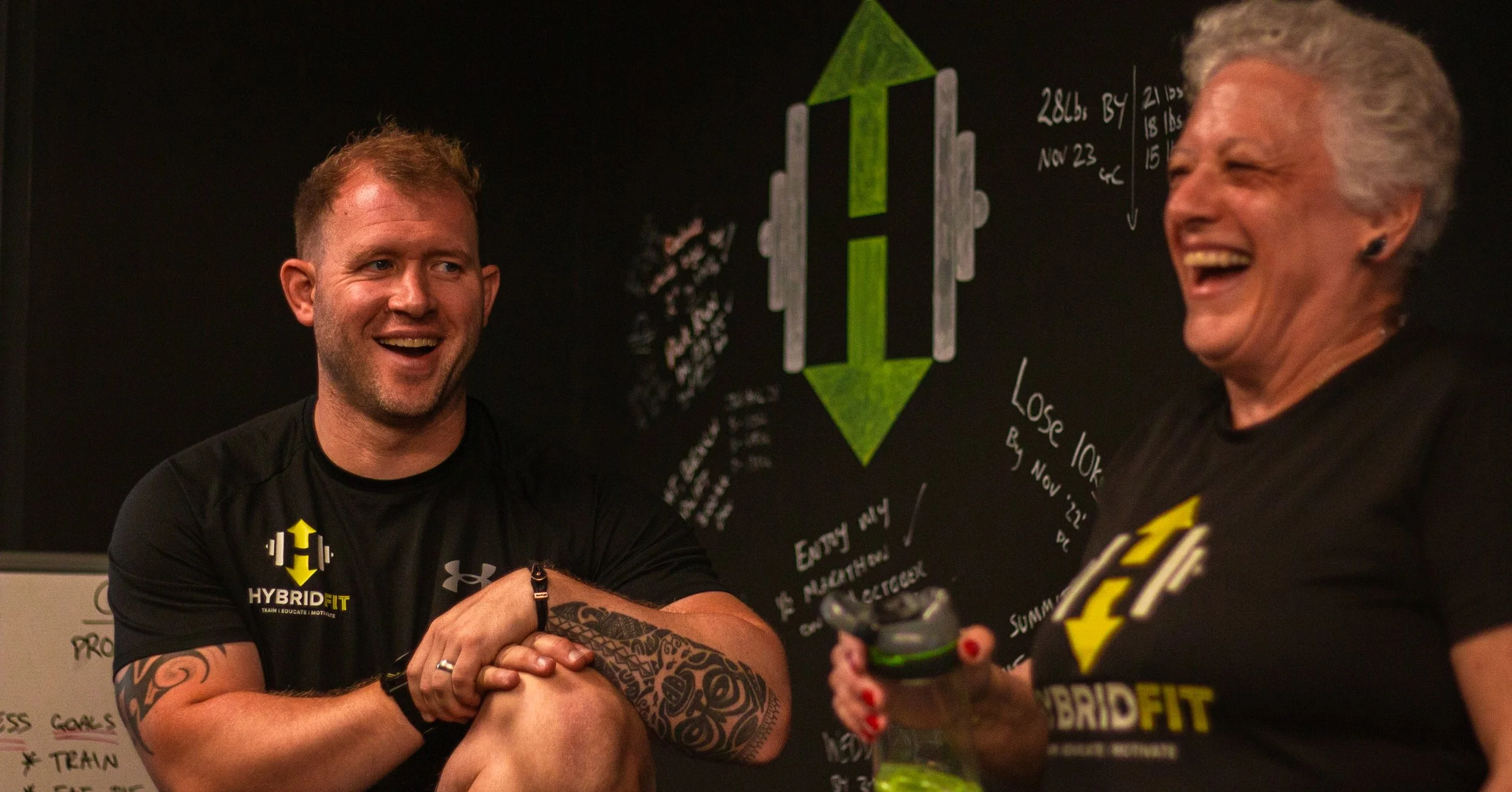 Two smiling people, a man and a woman, in workout shirts with 'HYBRID FIT' logos, are talking and laughing in a gym or fitness studio with a blackboard in the background that has diagrams and notes.