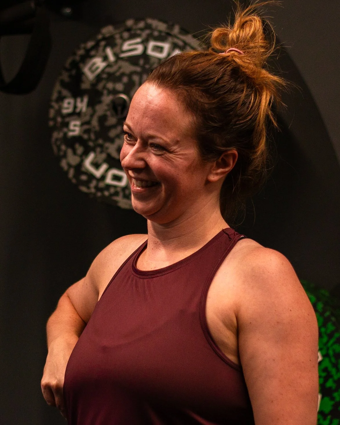 A woman with red hair tied in a bun, smiling and wearing a maroon athletic tank top, standing in front of a black background with a logo or design.