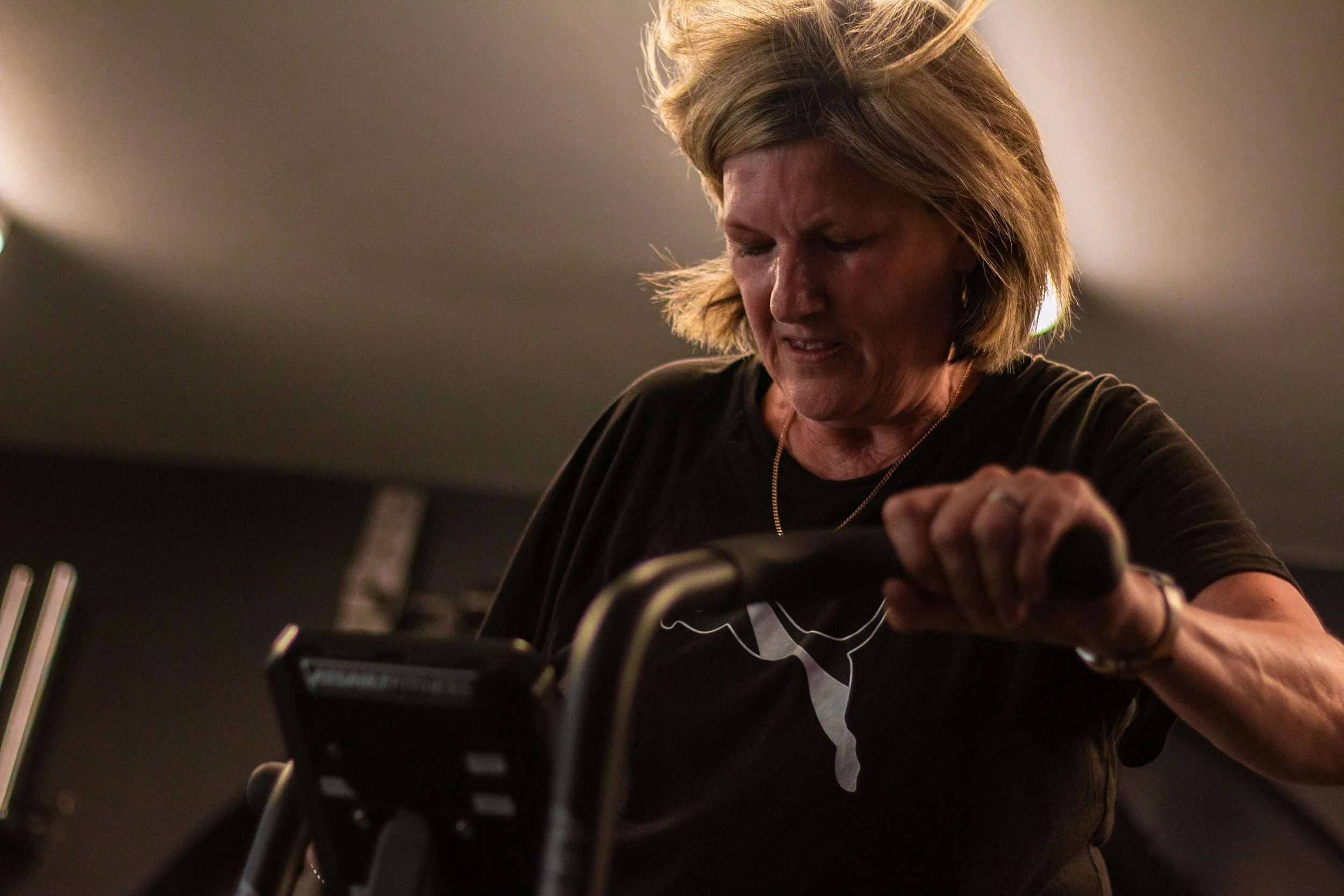 A woman with blonde hair looks down and grimaces as she grips the handlebar of a wheelbarrow or similar equipment.