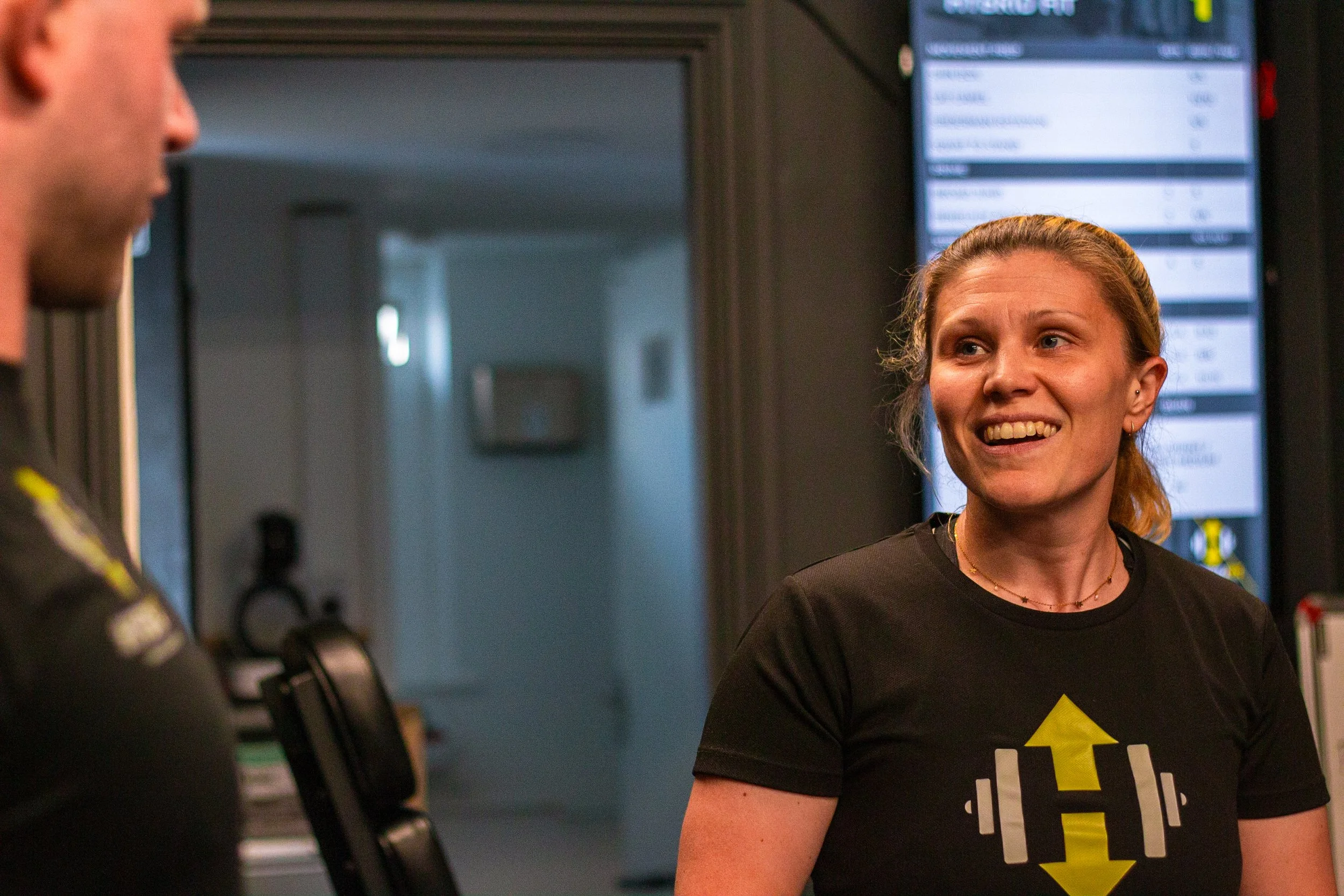 A woman smiling and talking to a man at a gym with a digital display board in the background.