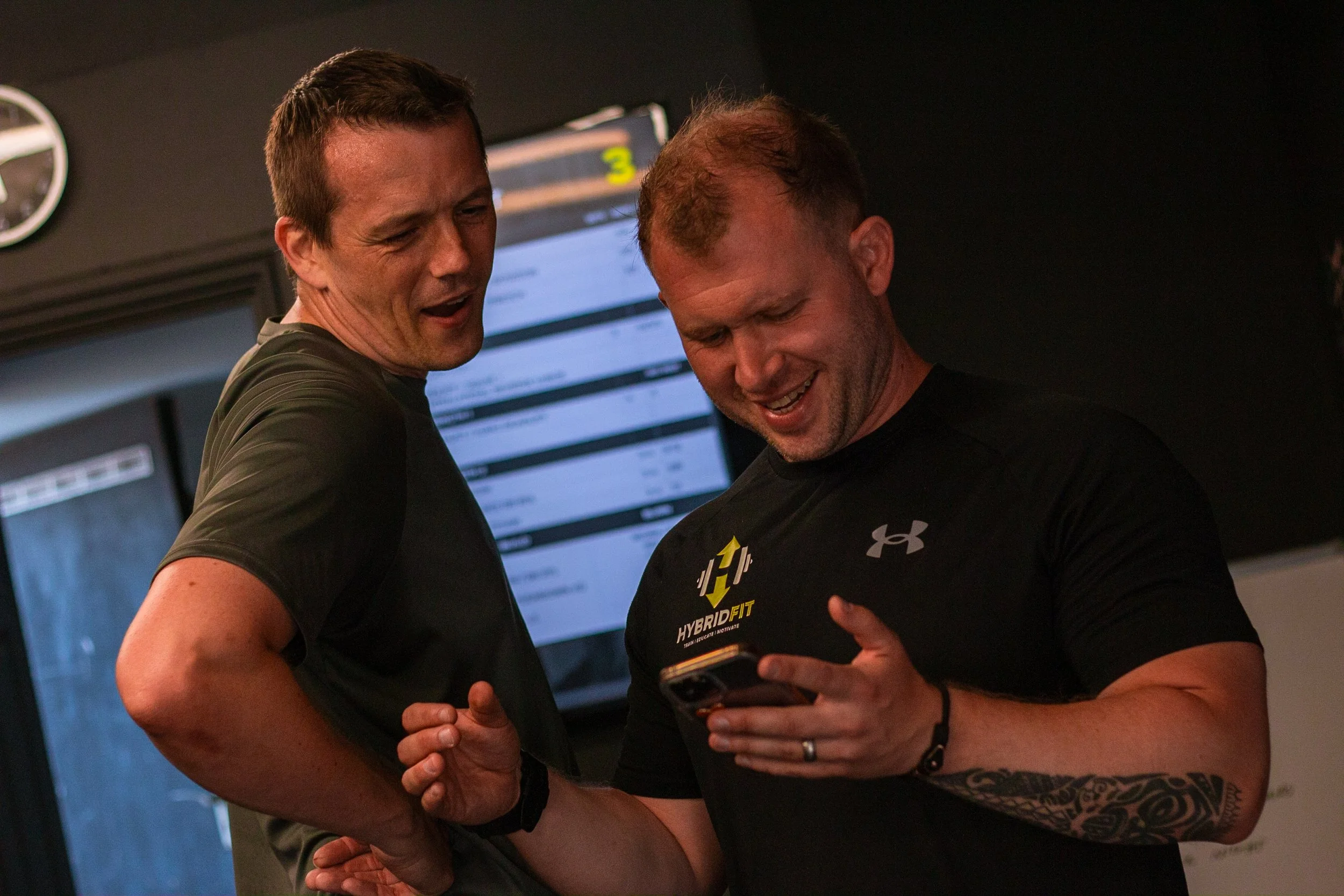 Two men smiling and looking at a smartphone in a gym setting, with a digital screen in the background.