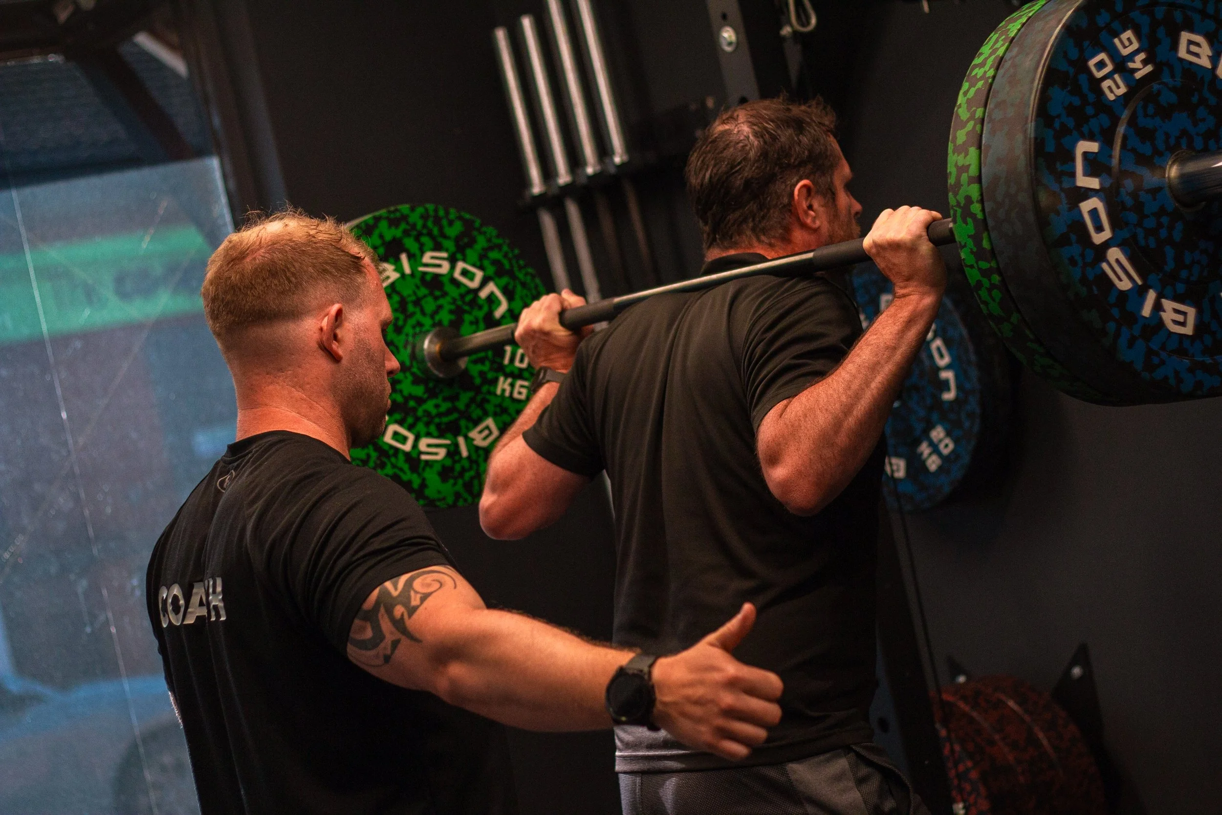Two men in black shirts working out at a gym; one is spotting the other who is lifting a barbell with weights on the wall.