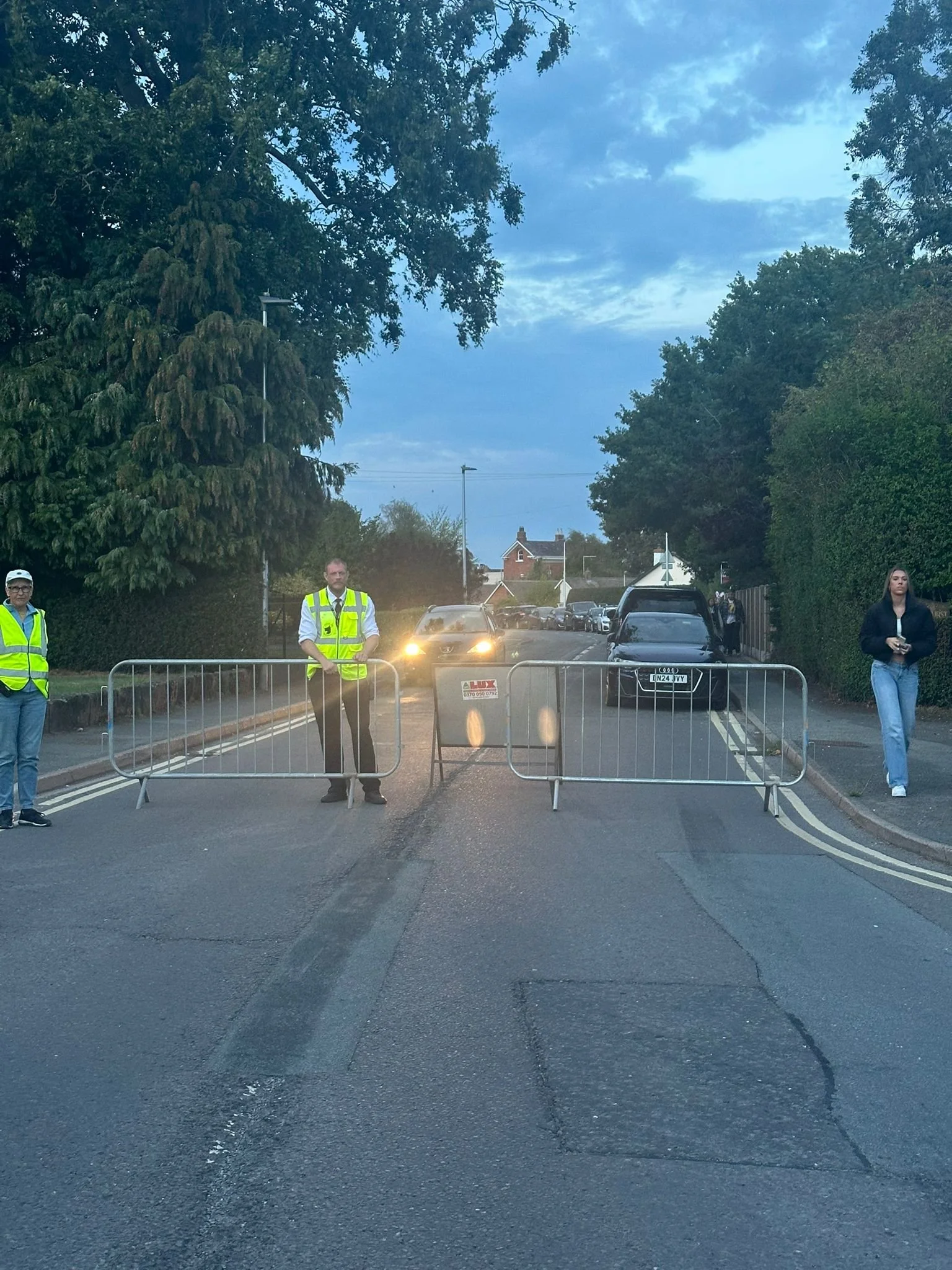 Street blocked by barriers with three people standing nearby. Vehicles in the distance with headlights on, trees lining the street, and a house visible further back.