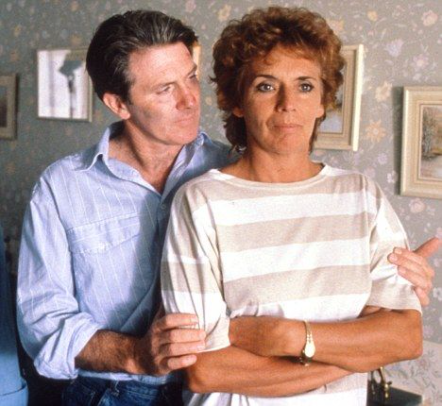 A man standing behind a woman, gently holding her arm, both looking serious and standing in a room with floral wallpaper and framed pictures.