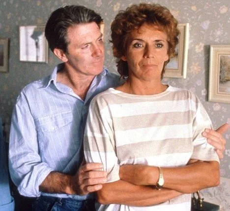 A man standing behind a woman, touching her arm, with a neutral expression, in a room with framed pictures on the wall.
