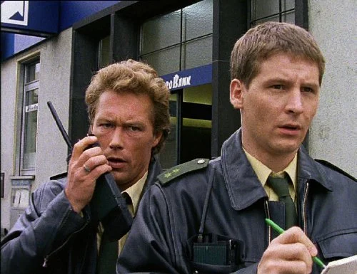 Two men in police uniforms outside a building, one using a walkie-talkie and the other holding a green pen.