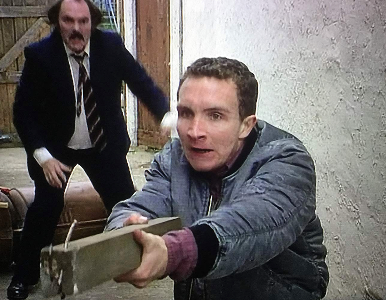 Two men in a barn or garage, one in the foreground holding an axe with a surprised expression, the other in the background reaching out with a surprised or aggressive look.