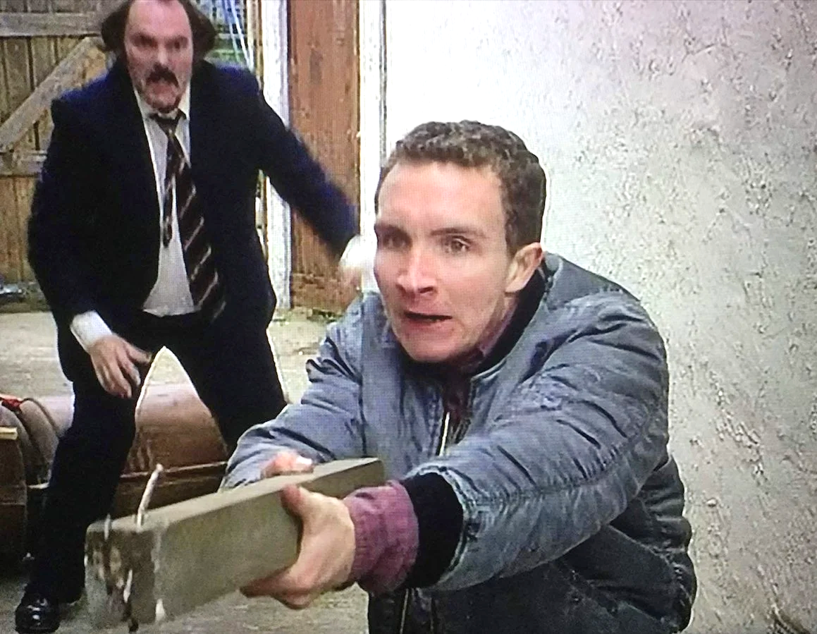 Two men inside a rustic barn, with one man in a leather jacket and dark hair holding a large wooden baseball bat, reaching toward the camera, while the other man with disheveled hair in a suit and striped tie is retreating.