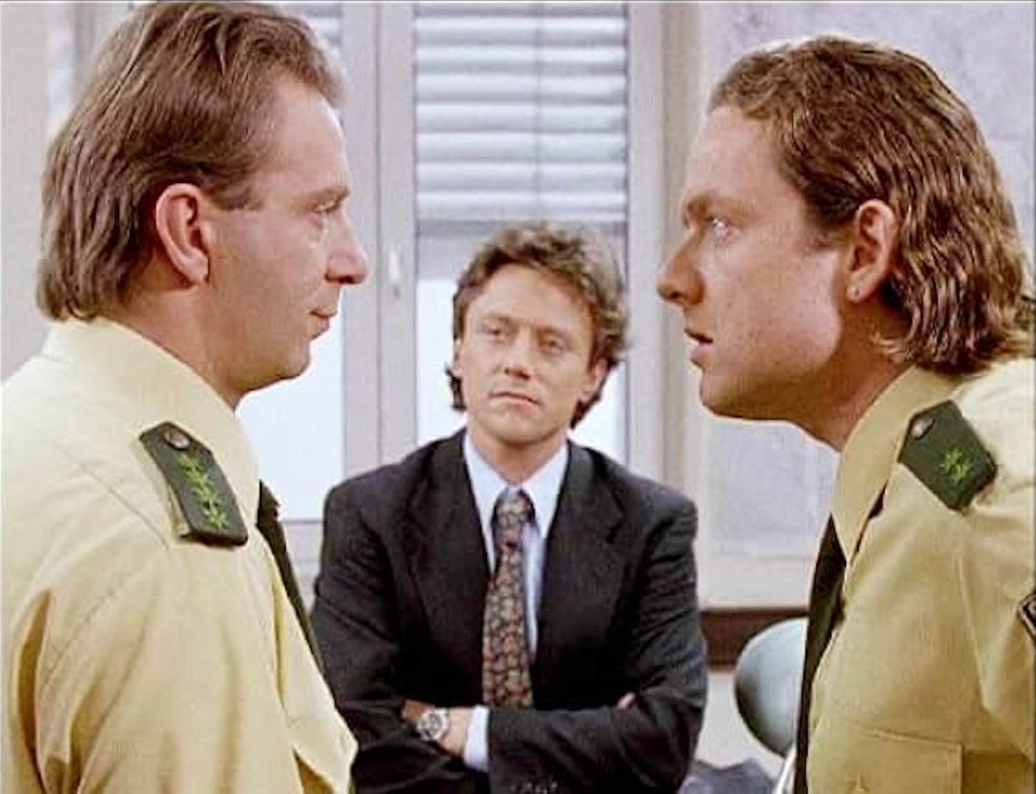 Two men in yellow military uniforms facing each other with serious expressions, and a man in a dark suit and patterned tie observing between them, in an office setting.