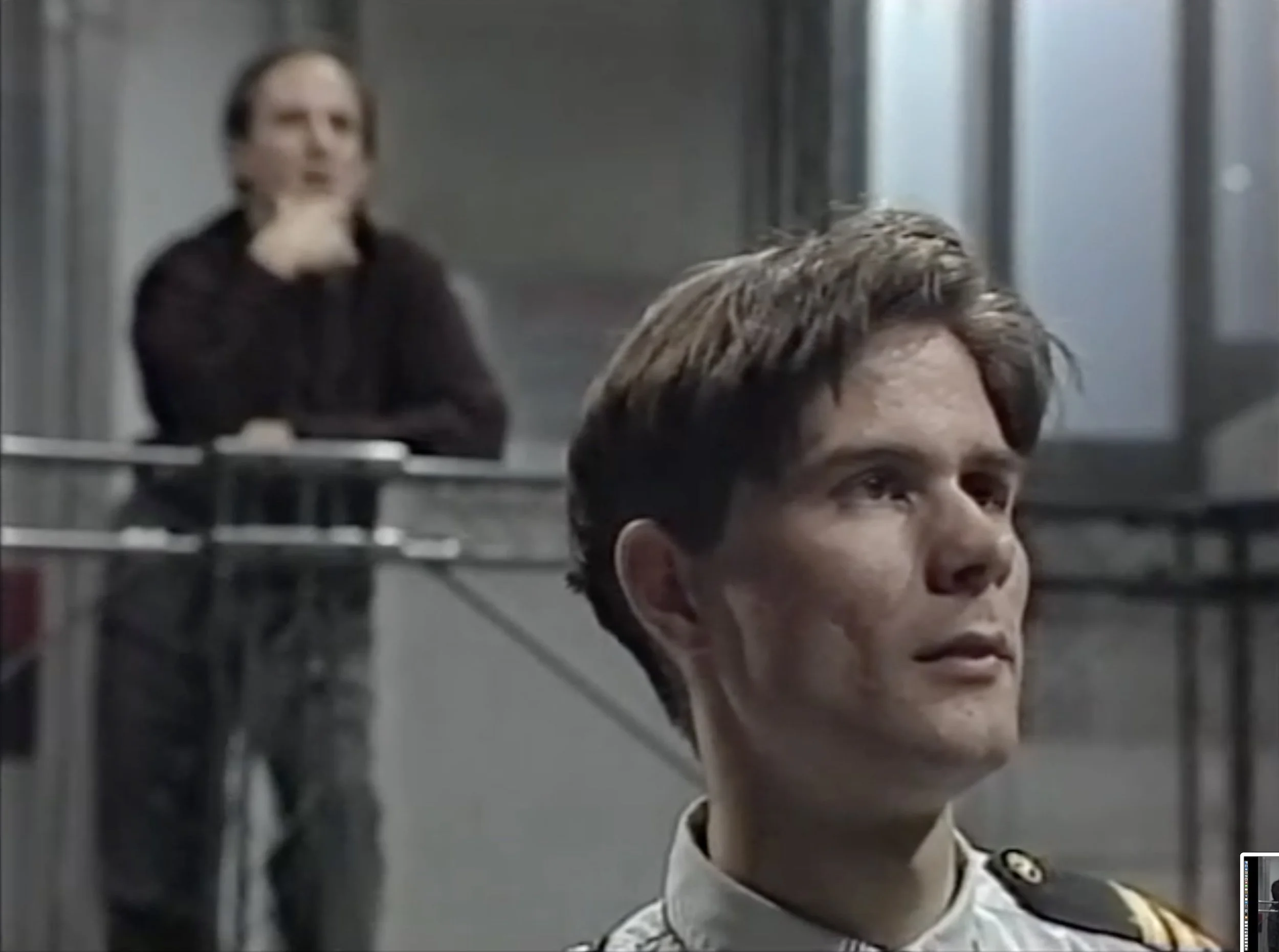 A young man in a military uniform with a slight smile in the foreground, a woman in black standing behind a glass barrier in the background, with her hand on her chin.
