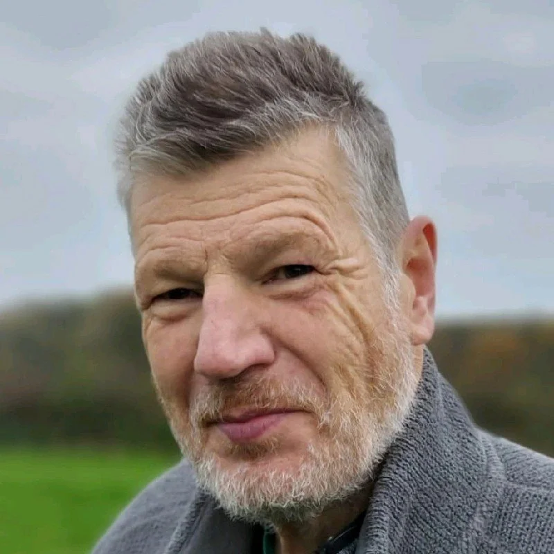 Close-up of a mature man outdoors, with short gray hair and a beard, smiling slightly, wearing a gray sweater, with a blurred green landscape background.