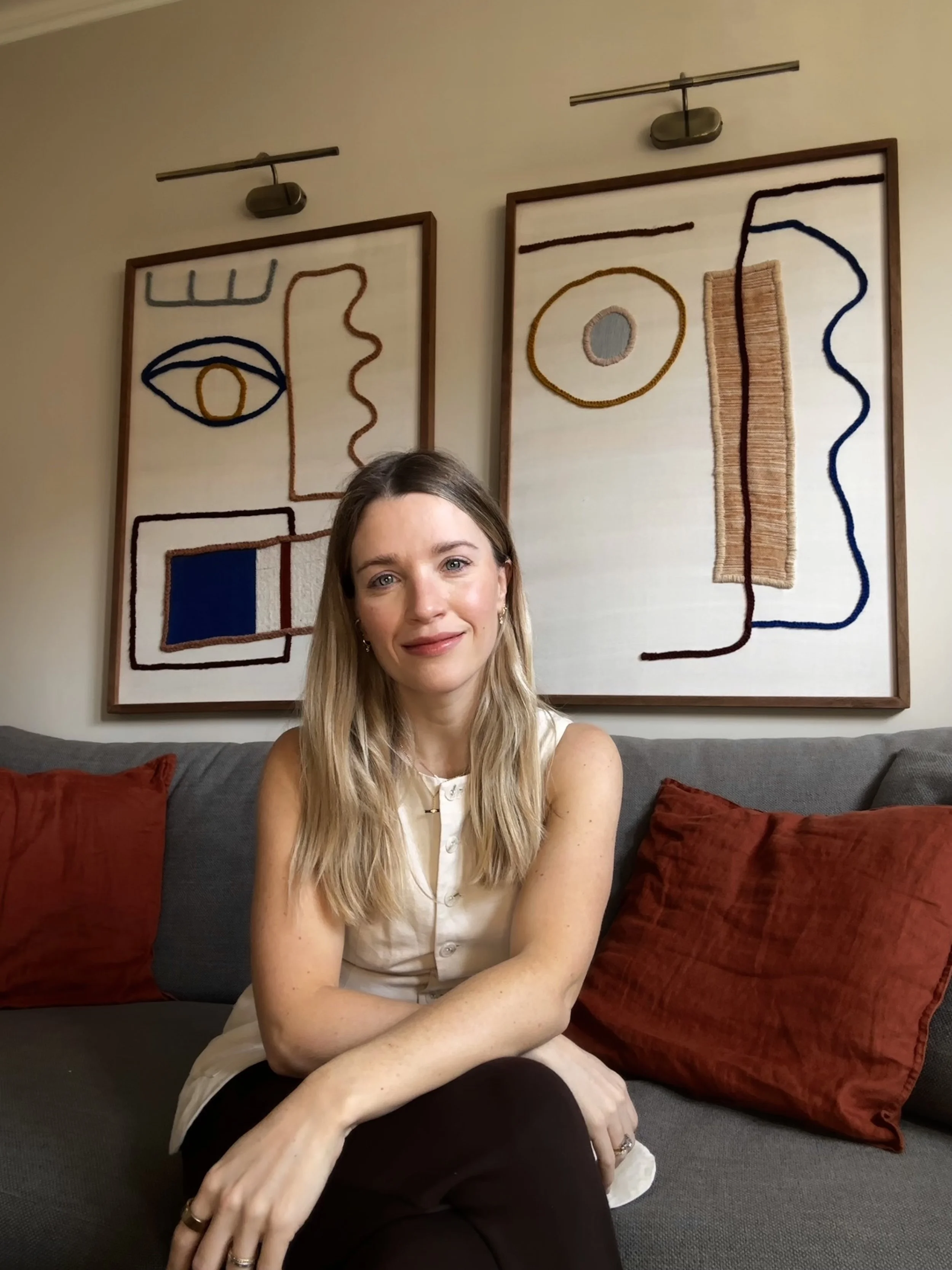 Sarah Keeble is sitting on a gray couch with red pillows, in front of abstract line artwork on the wall. Sarah has long blonde hair and is wearing a cream waistcoat.